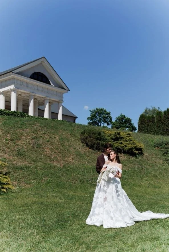 A bride and groom standing on a grassy hill in front of a large, white, Greek-style building with columns and a triangular pediment on a clear, sunny day.
