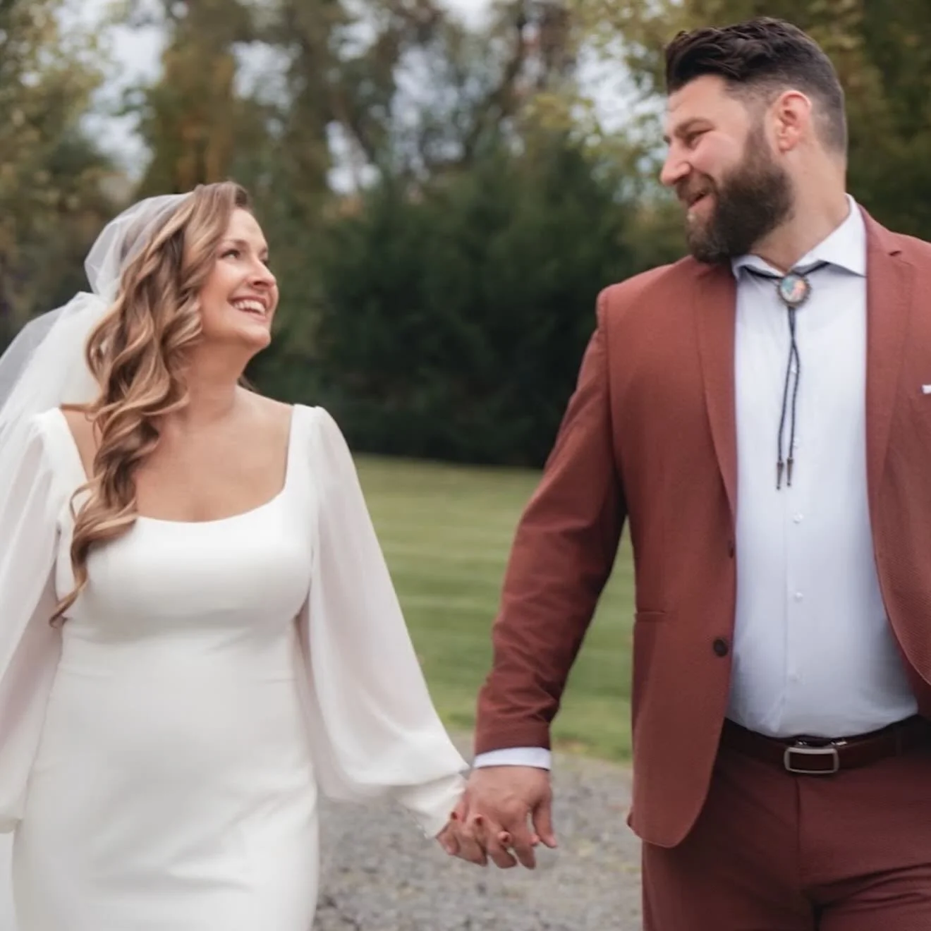 A bride and groom holding hands and smiling at each other outdoors on a grassy area with trees in the background, during a wedding photoshoot.