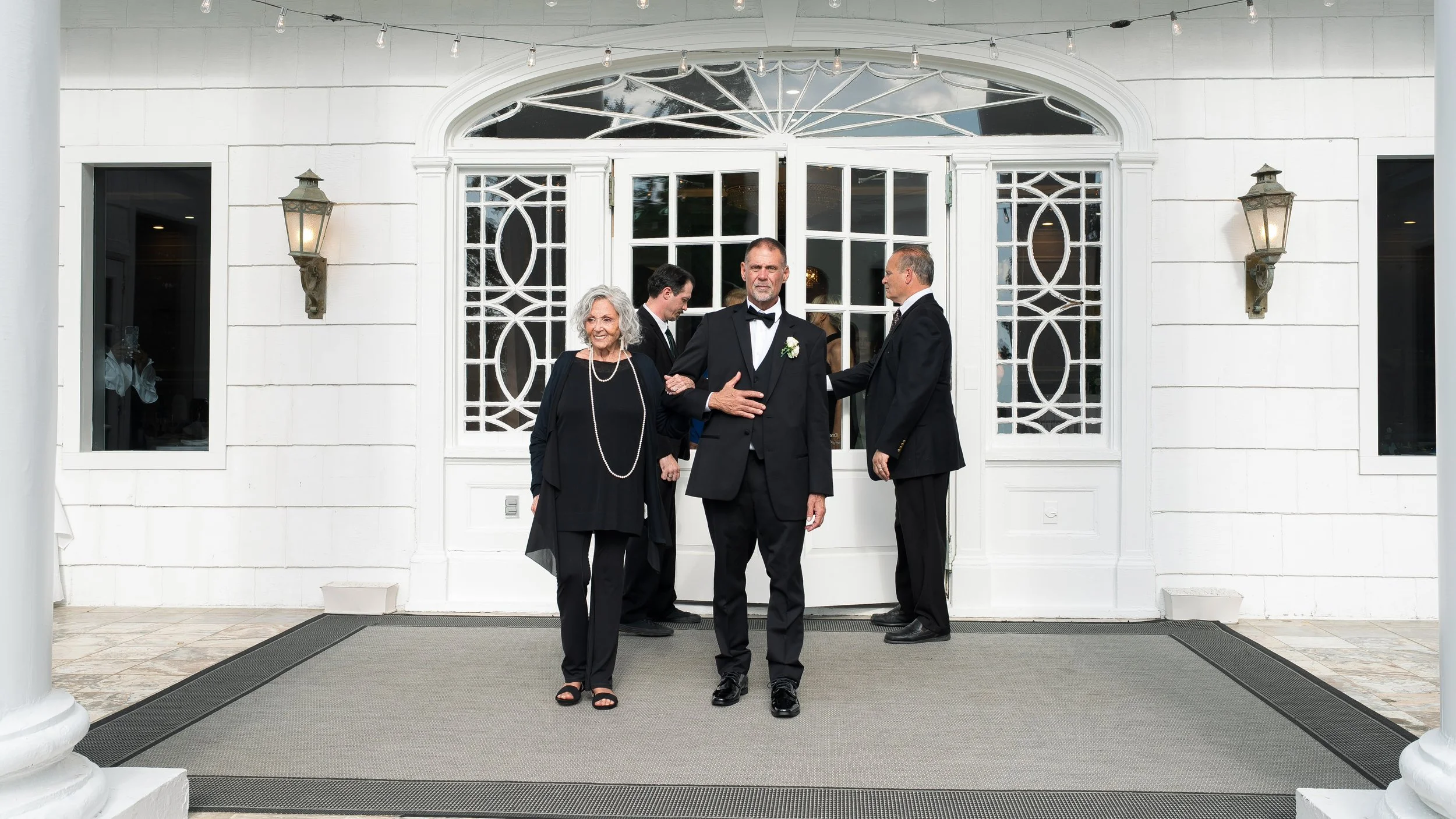 Three men in tuxedos and a woman in black dress standing outside of a white building with ornate glass doors, two wall-mounted lanterns, and string lights overhead.