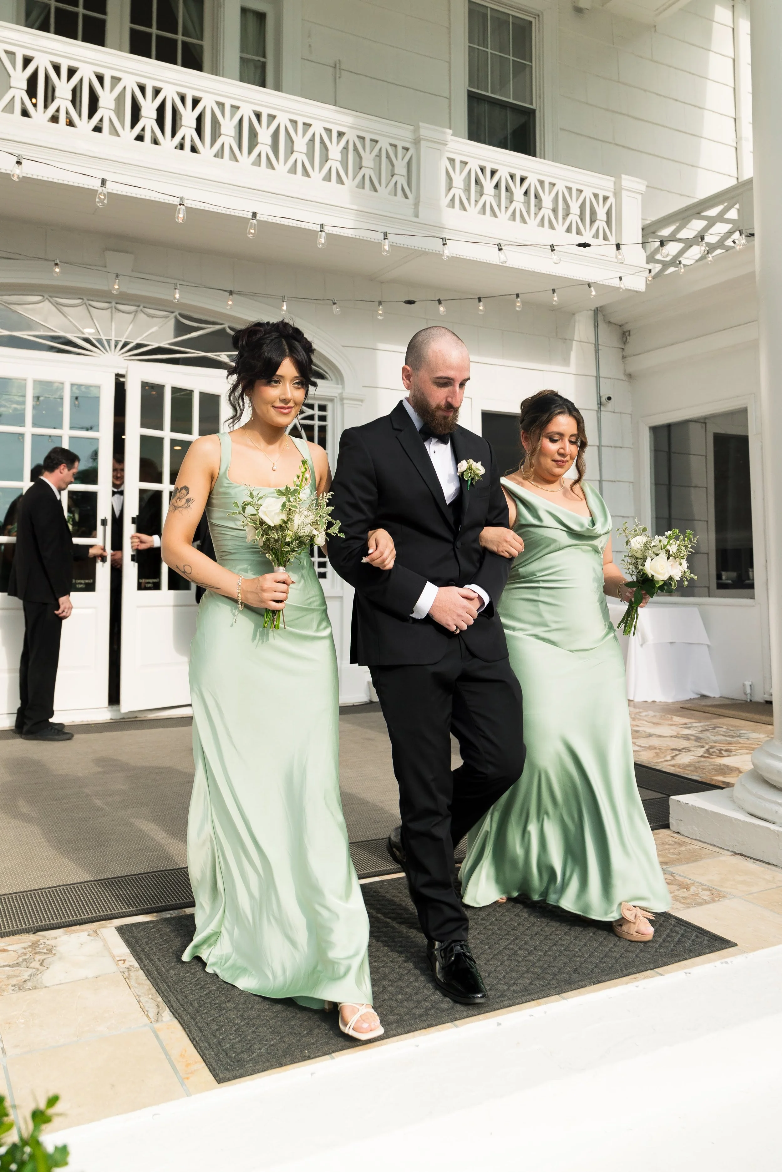 People in formal attire walking down an aisle at a wedding or event, with a woman in a light green dress holding a bouquet, a man in a black suit with a white shirt and black bow tie, and another woman in a matching light green dress holding a bouque