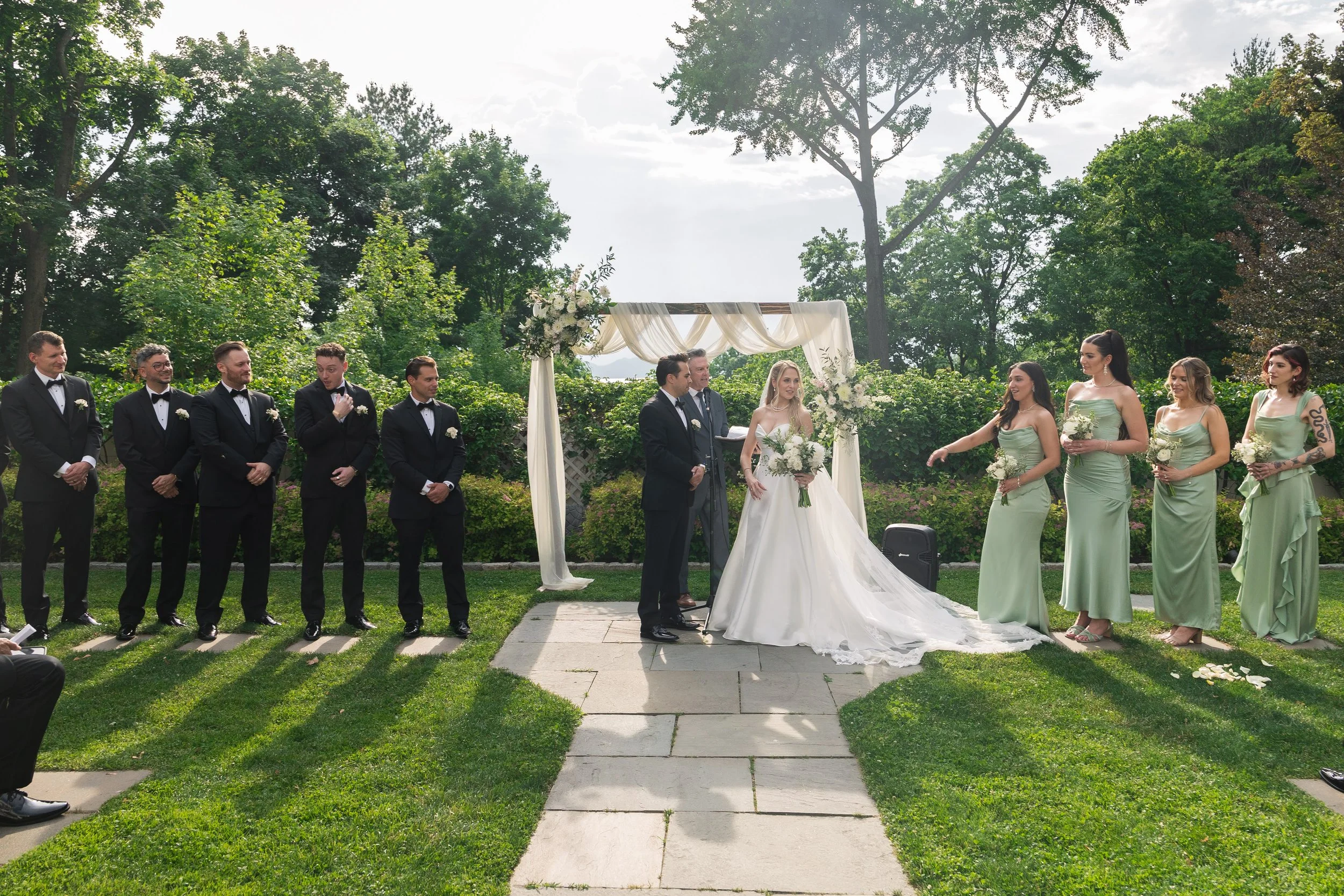 Outdoor wedding ceremony with the bride, groom and wedding party standing under a decorated arch in a garden setting with trees and cloudy sky in the background.