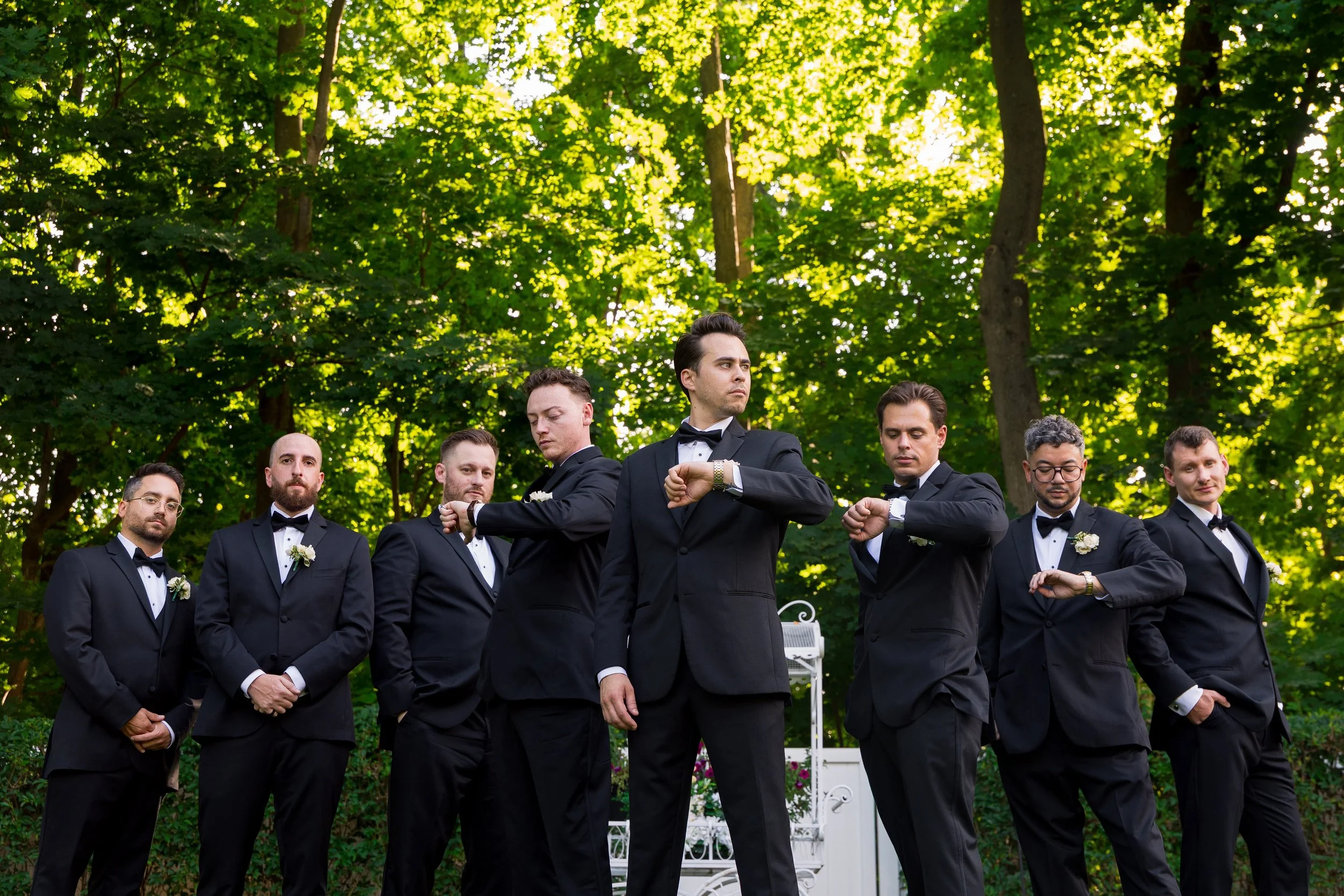 A group of men in tuxedos standing outdoors among green trees, with one man looking at his watch in the center.