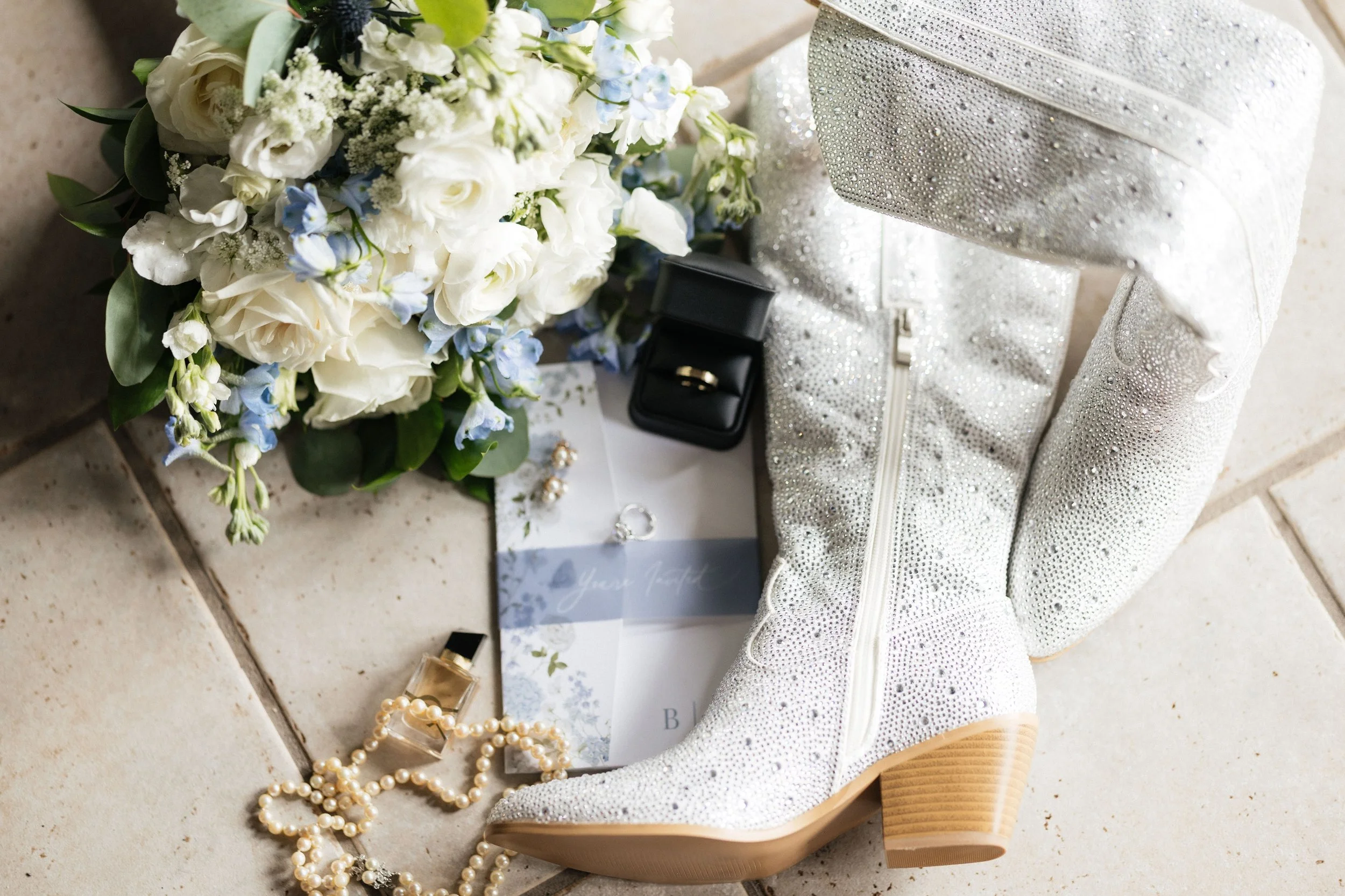 A collection of wedding accessories on a tiled surface, including a white Minnie Mouse-style shoe with sparkling details, a bouquet of white and light blue flowers, a black jewelry box with a ring inside, a string of pearls, and a wedding invitation.