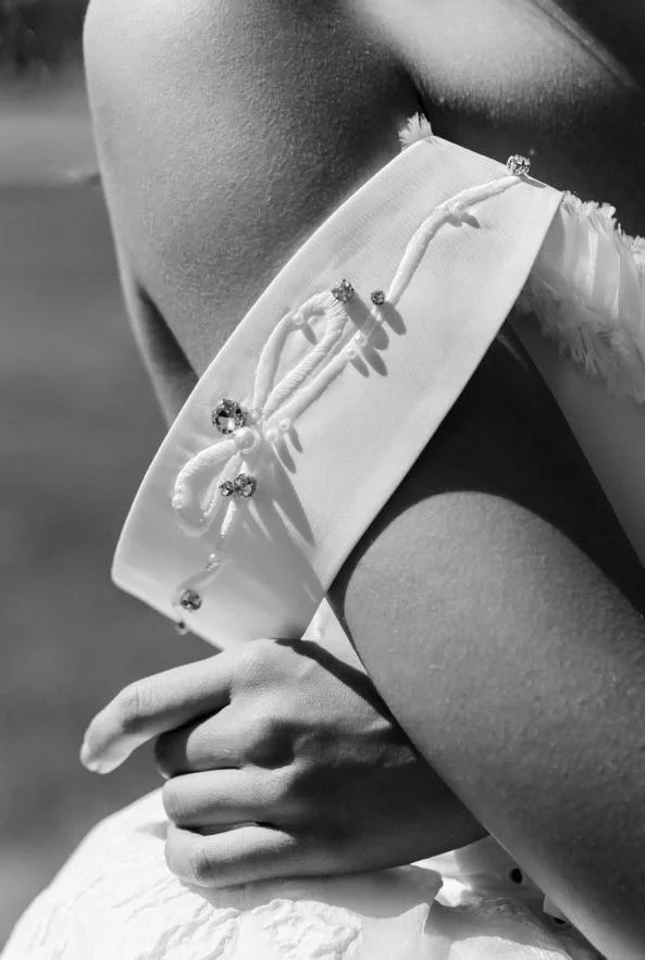 Close-up black and white photo of a woman wearing a sleeveless dress with rhinestone embellishments, holding a white clutch purse with a bow detail, outdoors with a blurred background.