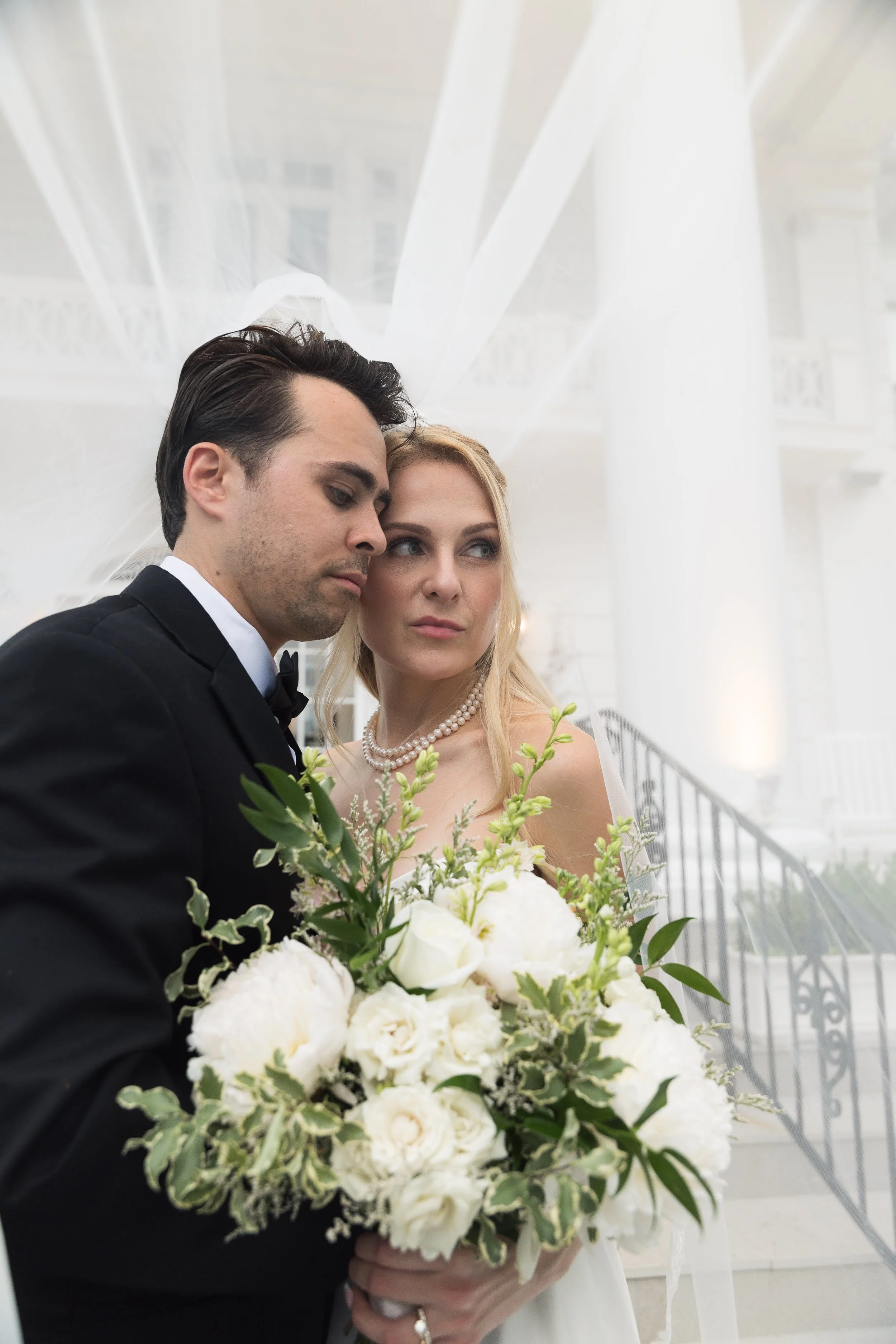 A bride and groom at their wedding, holding a bouquet of white flowers, standing close to each other in a white, elegant venue.