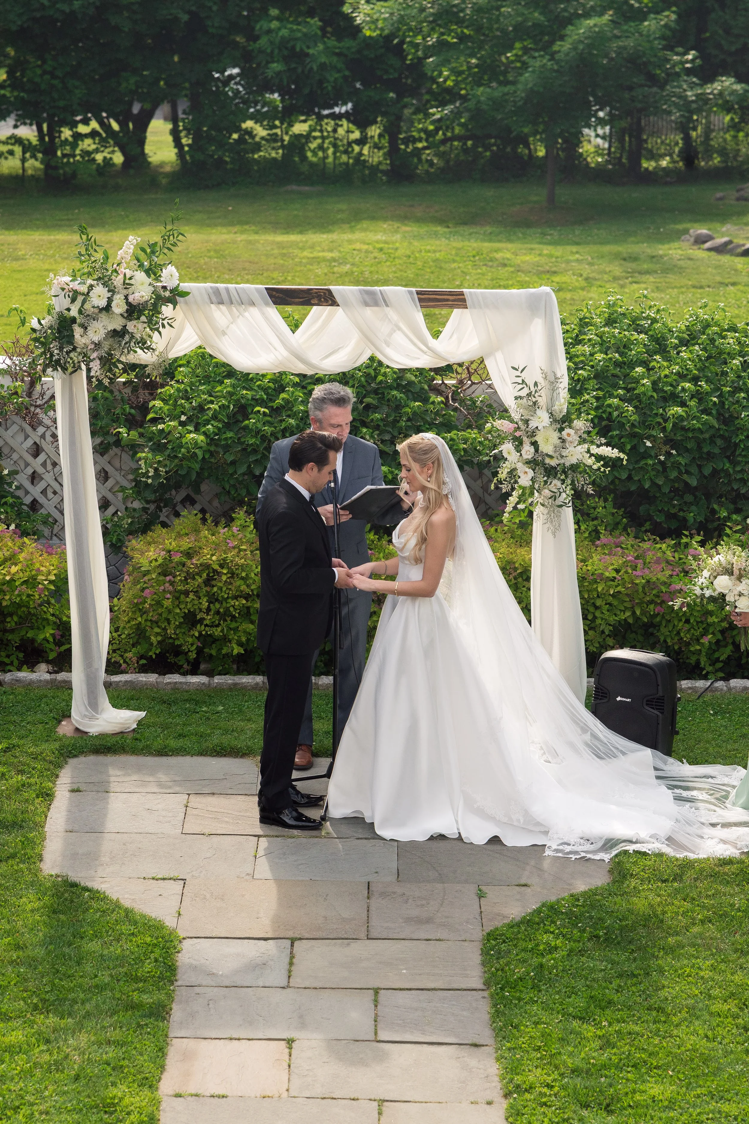 A bride and groom holding hands during their wedding ceremony outdoors beneath a white fabric-draped arch decorated with flowers. An officiant stands between them, reading from a book. Lush green bushes and trees surround the scene.