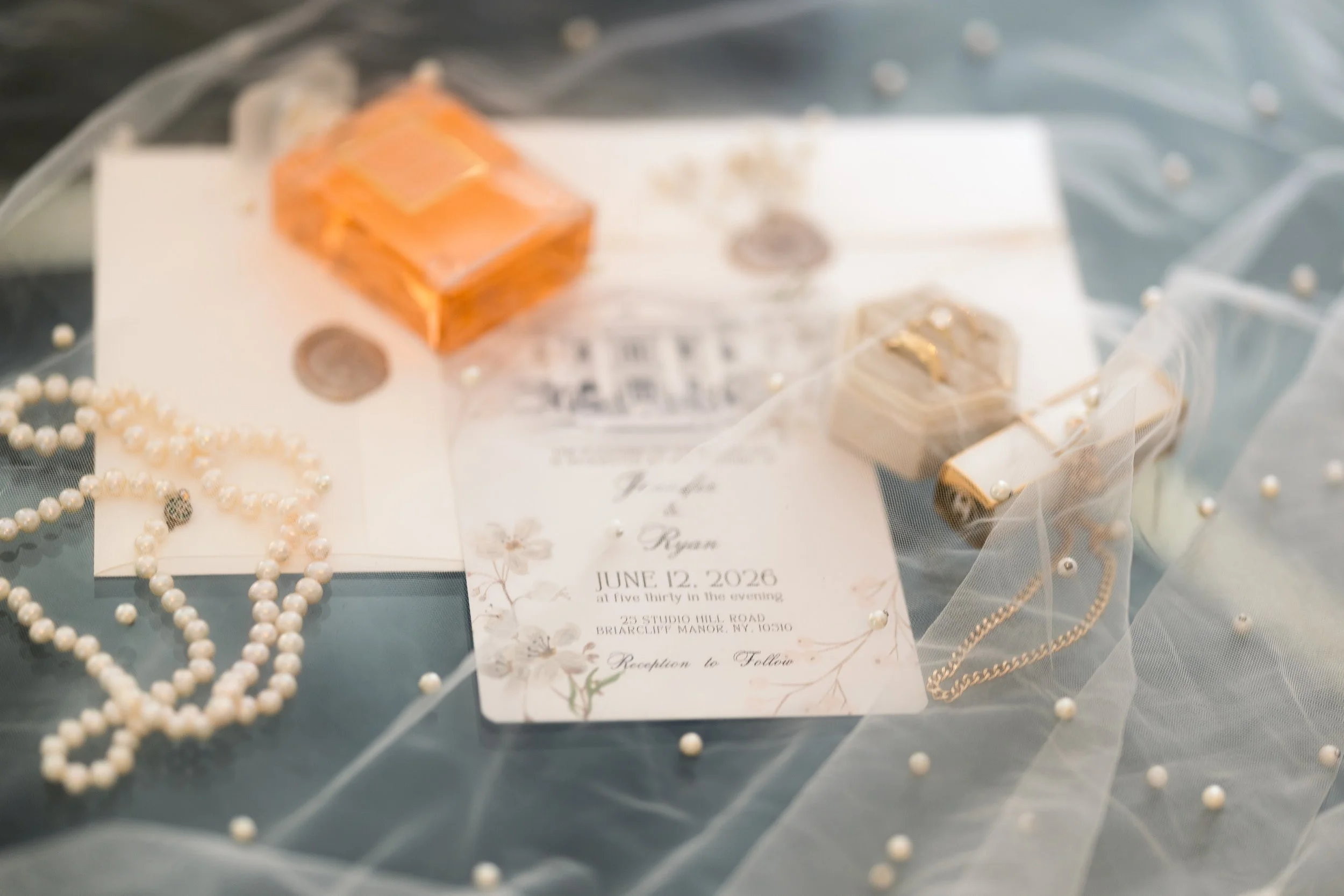 A collection of jewelry including pearl necklaces and gold chains, along with a wedding invitation for June 12, 2026, at Five Thirty in the evening, at 23 Studio Hill Road, Briarcliff Manor, NY. The jewelry is displayed on a sheer white fabric with s