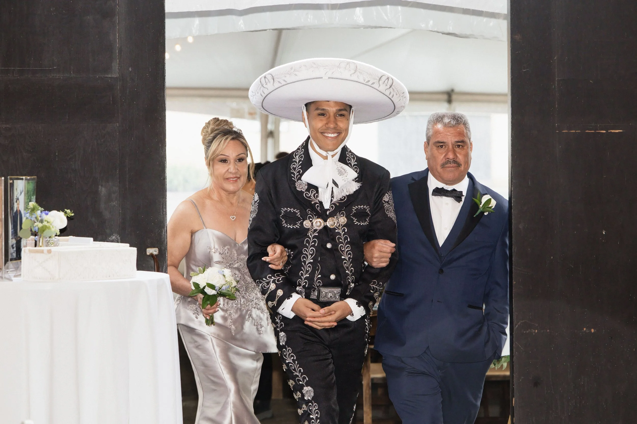 A young man dressed in traditional Mexican charro attire, including a wide sombrero, walks arm in arm with a woman in a white wedding dress who is holding a bouquet. They are flanked by an older man in a tuxedo, all smiling.
