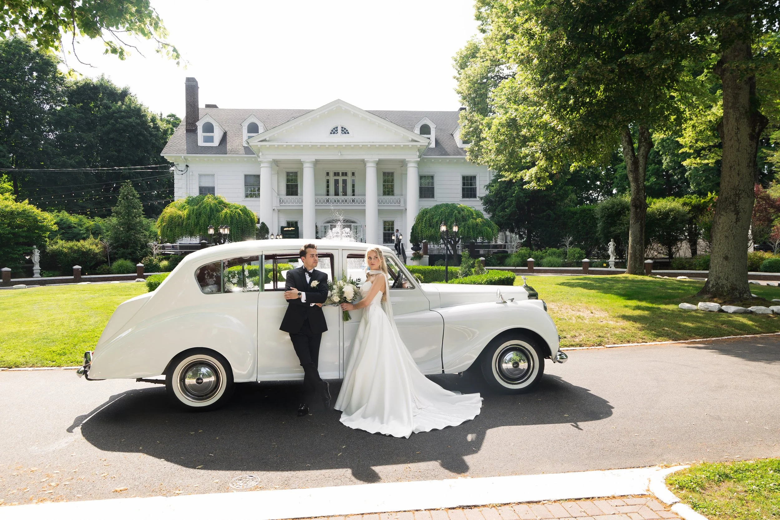 A bride and groom in wedding attire standing beside a vintage white car parked on the street in front of a large white house with a grand porch and lush green trees.