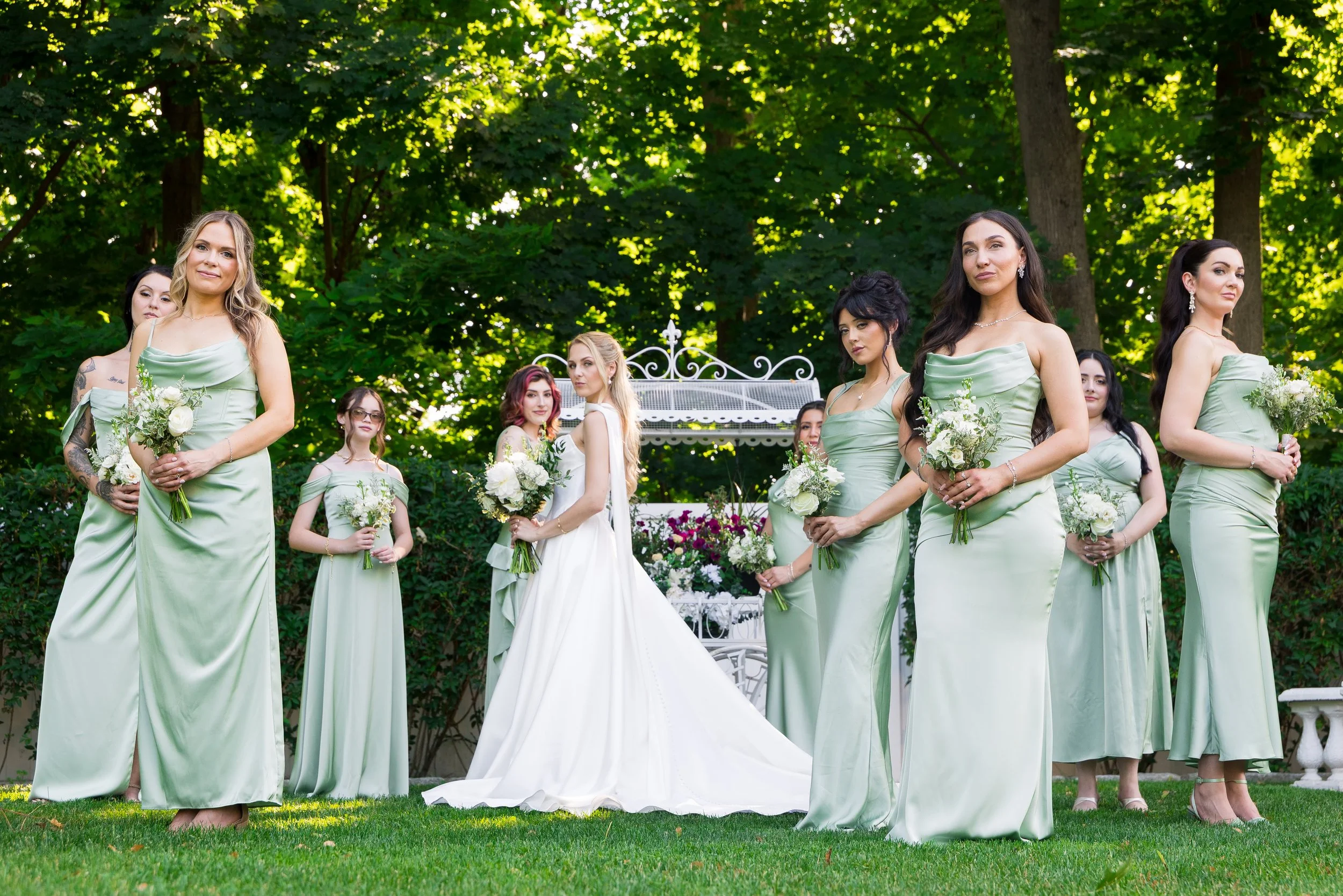 A group of women dressed in matching light green dresses, standing outdoors on grass, holding bouquets of white flowers, with trees and greenery in the background.