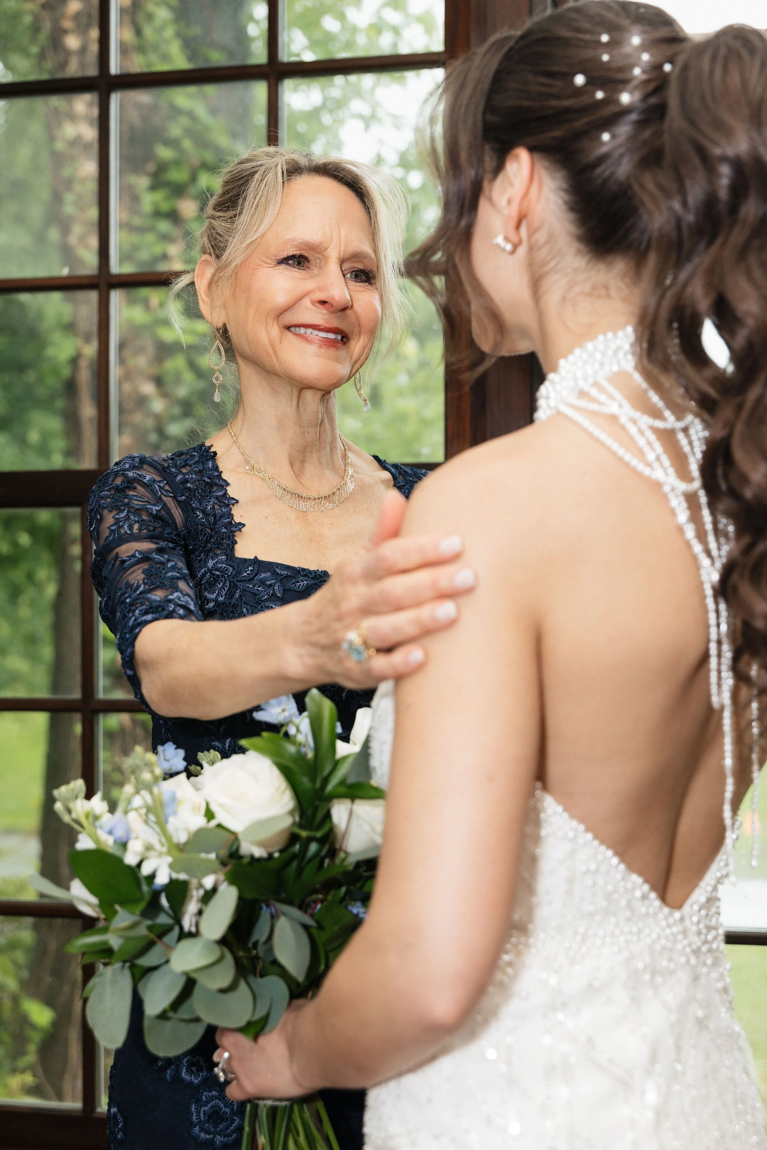 An emotional moment at a wedding as an older woman with blonde hair, wearing a navy lace dress and pearl earrings, embraces the bride, who is holding a bouquet of white and purple flowers. The bride, with brown curly hair adorned with pearls, is wear