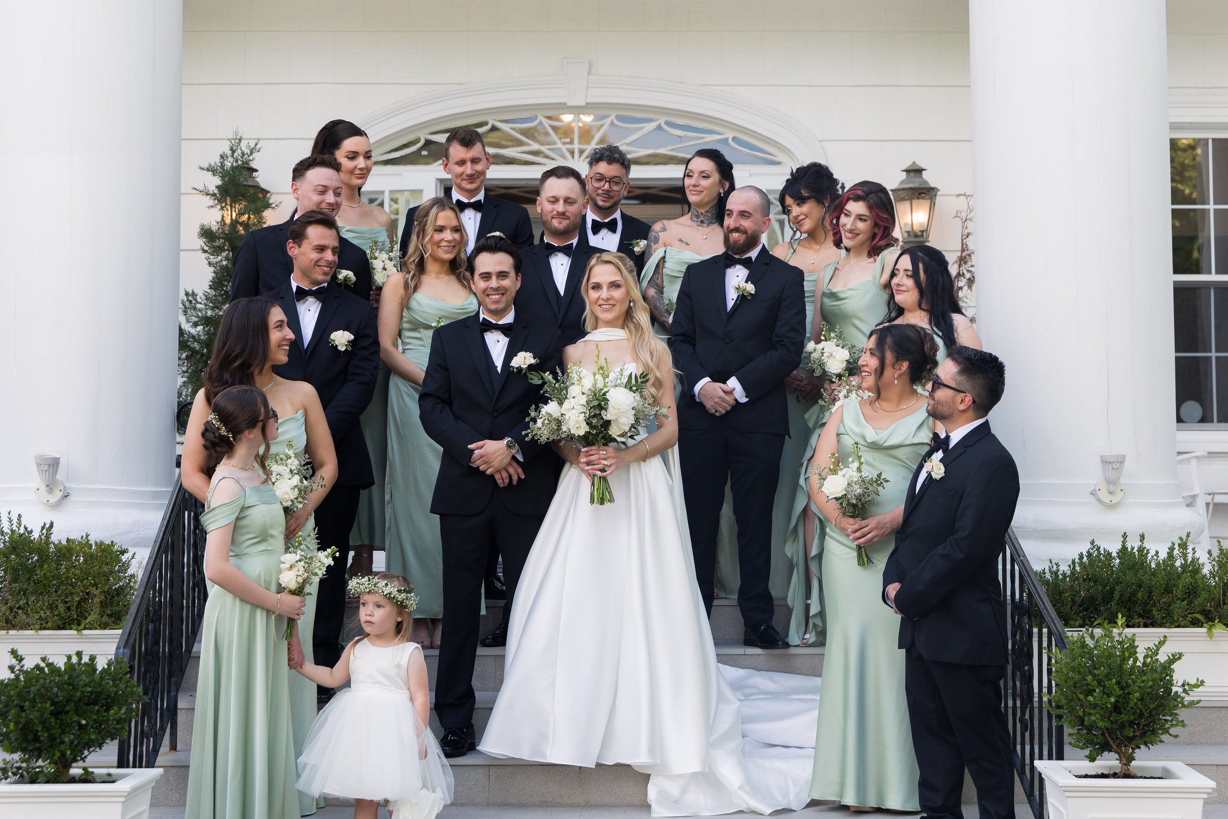 Group of people dressed in formal attire outside a building with white columns, celebrating a wedding. The bride is holding a bouquet of white flowers, and several bridesmaids and groomsmen surround her, smiling and posing for the photo.
