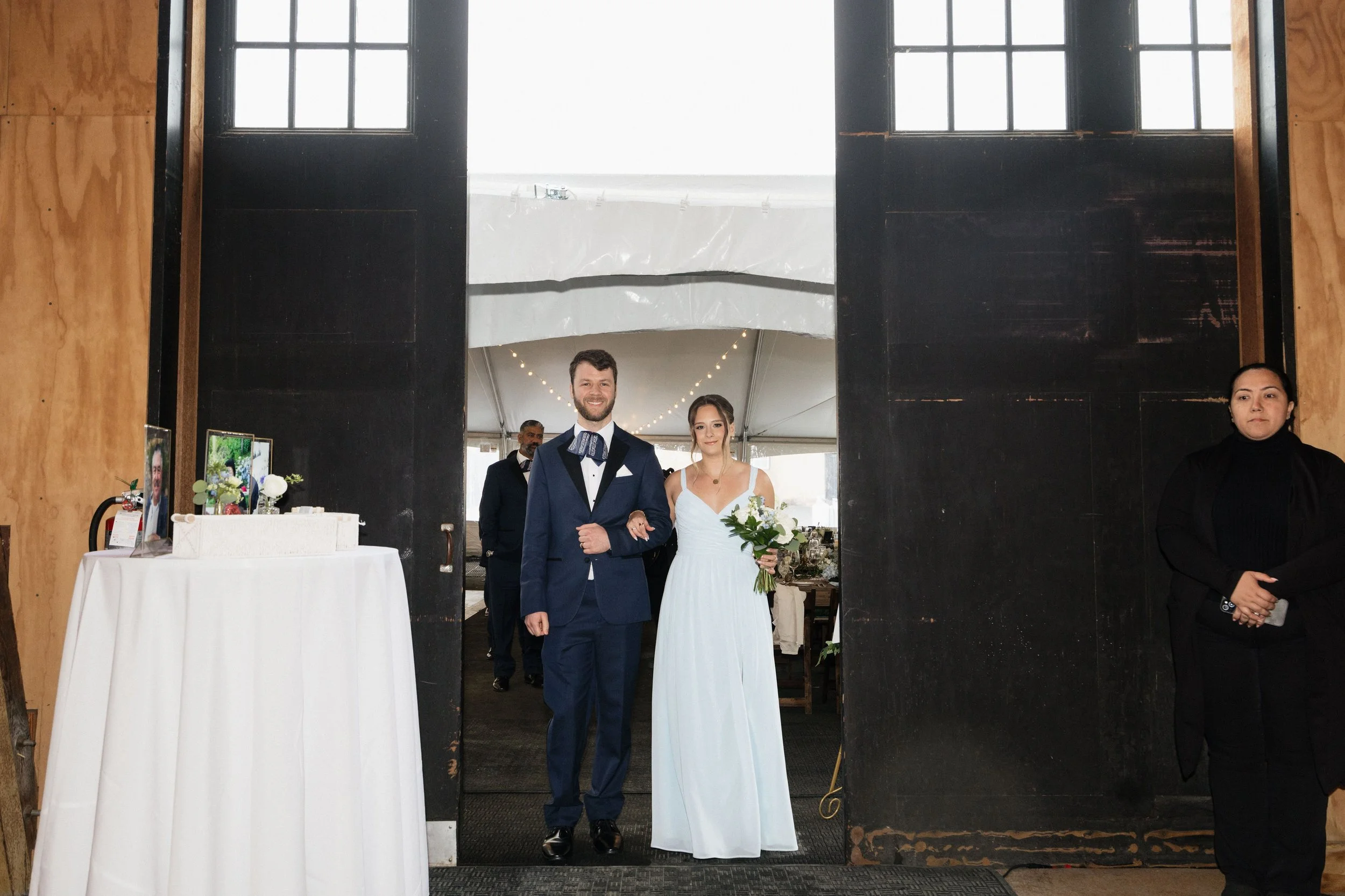 A bride and groom smiling as they walk out of a building, holding hands. The groom is in a navy tuxedo, and the bride is in a white wedding dress holding a bouquet of flowers. Behind them are guests and a decorated event space.