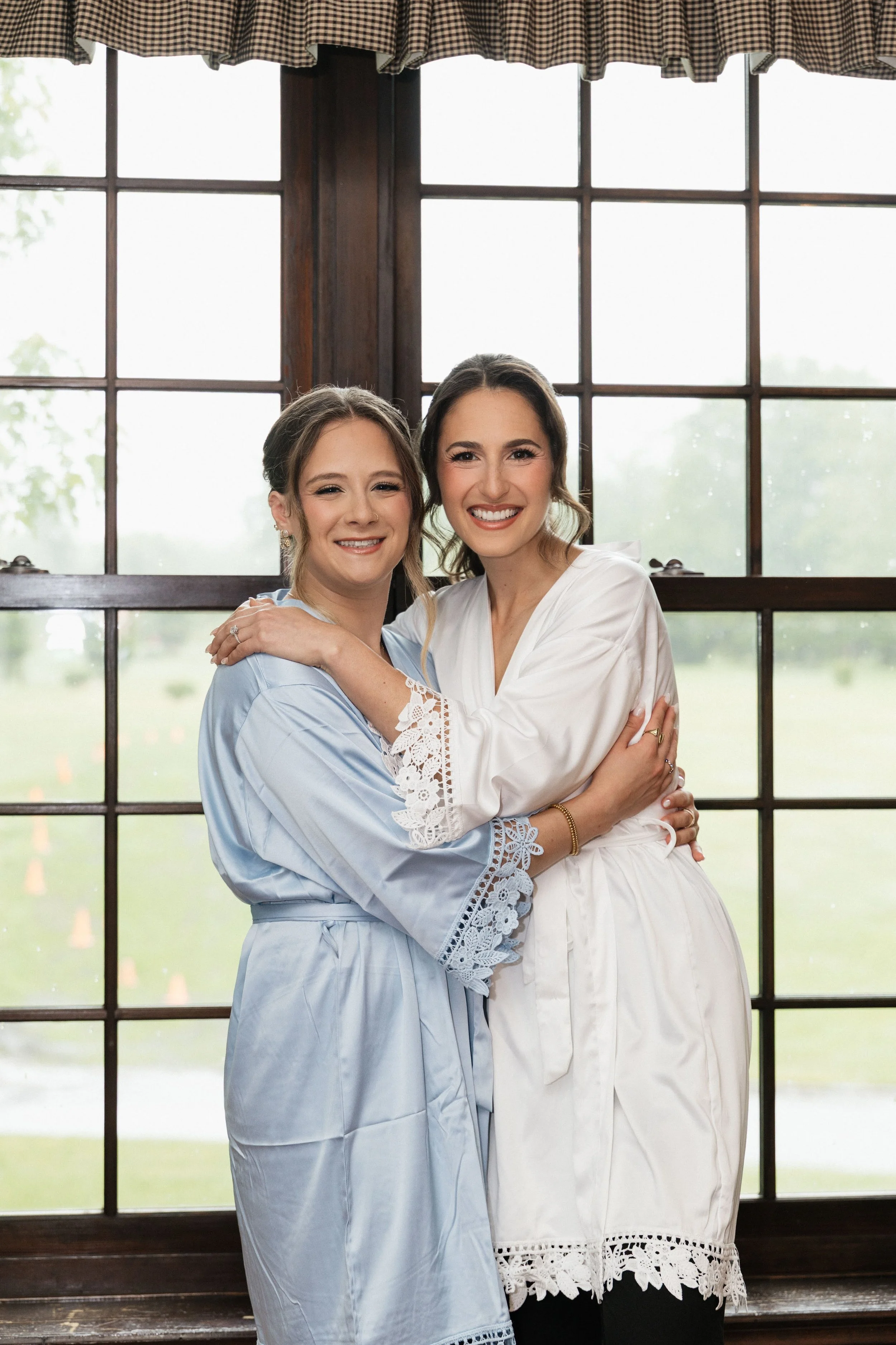 Two women smiling and hugging each other in front of a large window with a checkered valance, daytime.