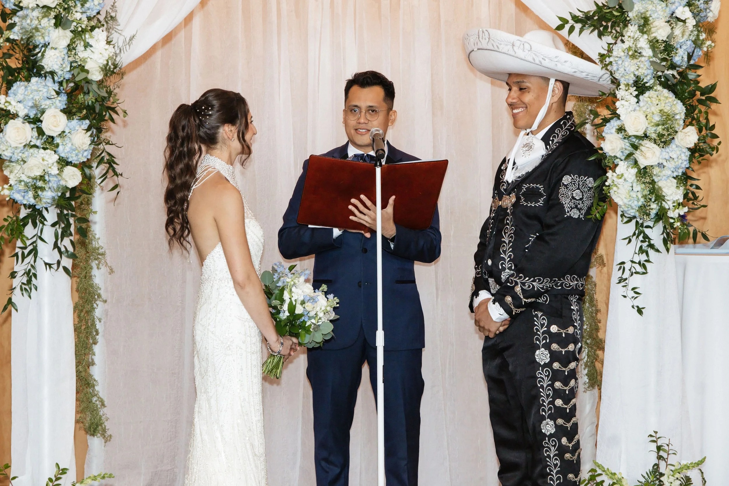 A wedding ceremony with a bride and groom exchanging vows, officiant, and decorated floral arch.