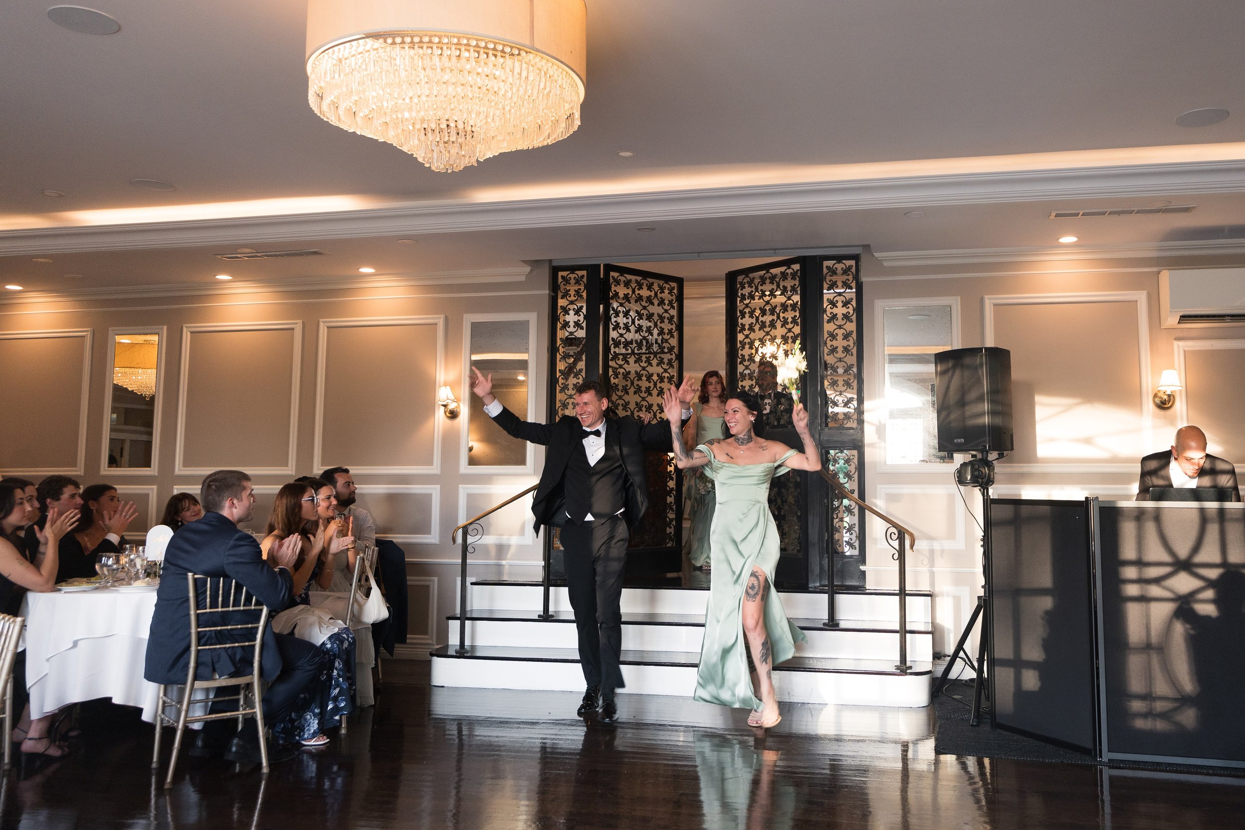 A wedding reception with a bride and groom dancing on a stage. The bride is in a green dress with tattoos on her leg, holding a flower, and the groom is in a black tuxedo. Guests are seated at tables clapping and watching. The room has elegant decor 