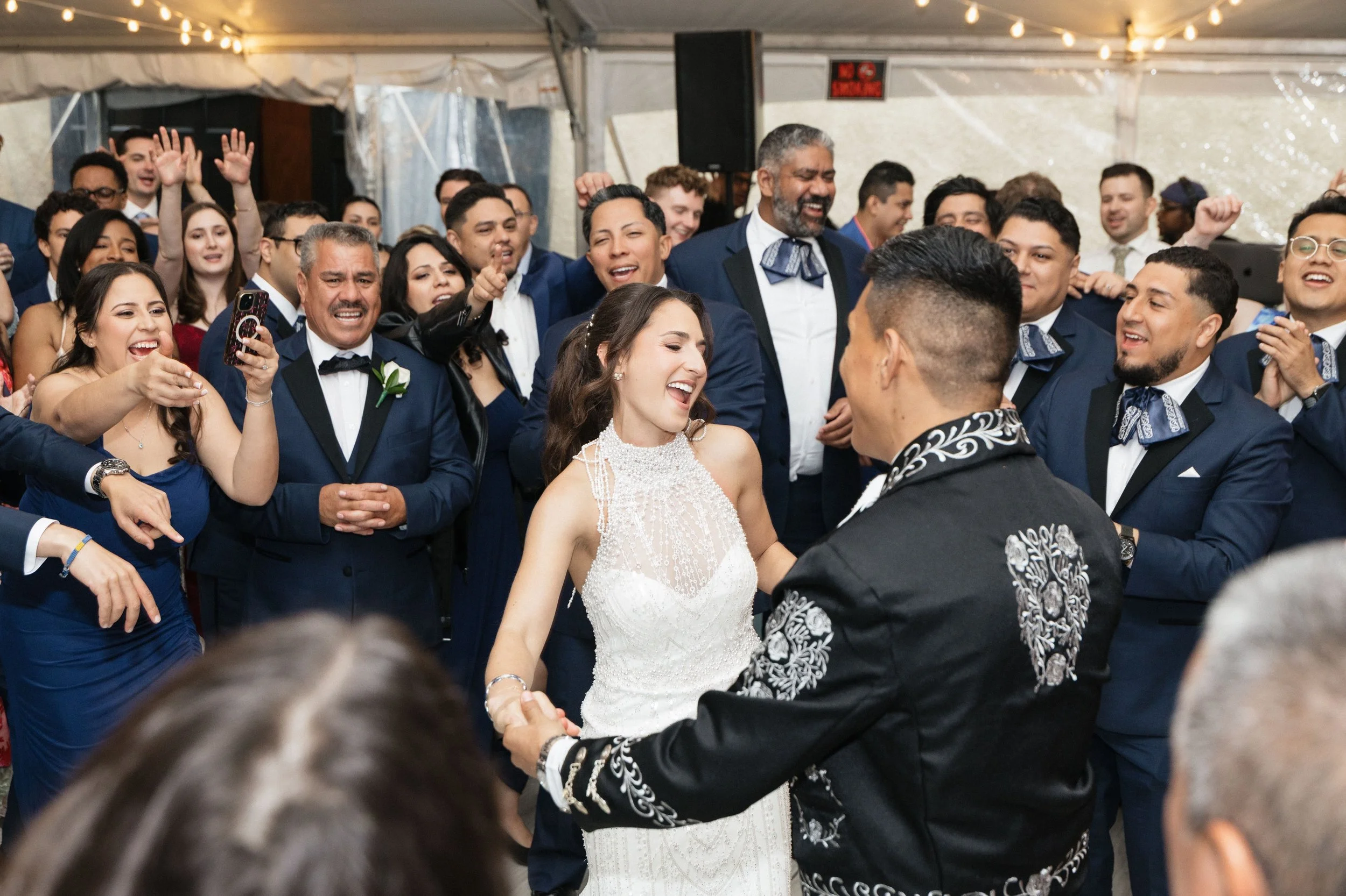 A group of people dressed in formal attire, including tuxedos and an elegant white dress, are celebrating at a wedding reception. The bride and groom are dancing and holding hands, surrounded by friends and family who are smiling, cheering, and captu