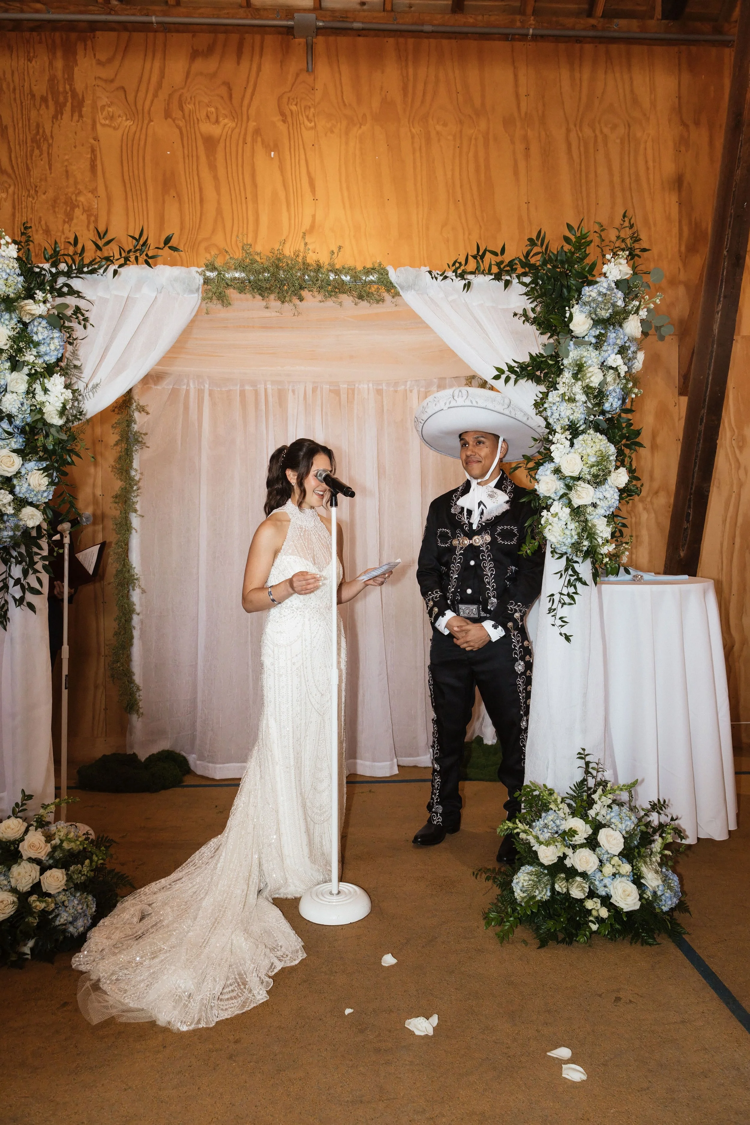 A bride and groom exchange vows under a floral arch at their wedding ceremony. The bride wears a white lace gown, and the groom is dressed in traditional mariachi attire with a wide-brimmed hat. Flowers and greenery decorate the setting.