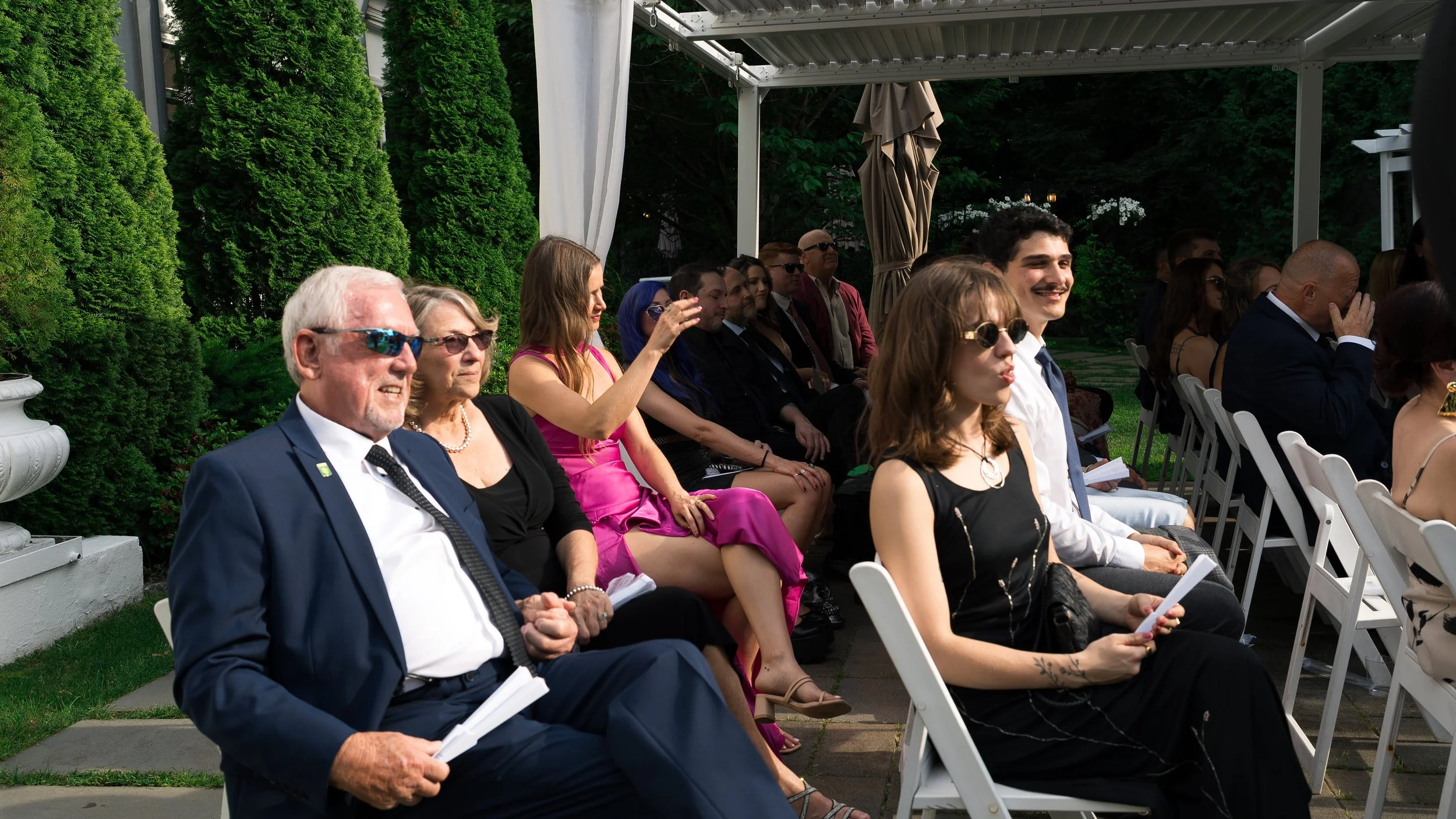 A group of people attending an outdoor event, seated on white chairs under a covered area with green trees in the background. The attendees are dressed in formal and semi-formal attire, some wearing sunglasses.