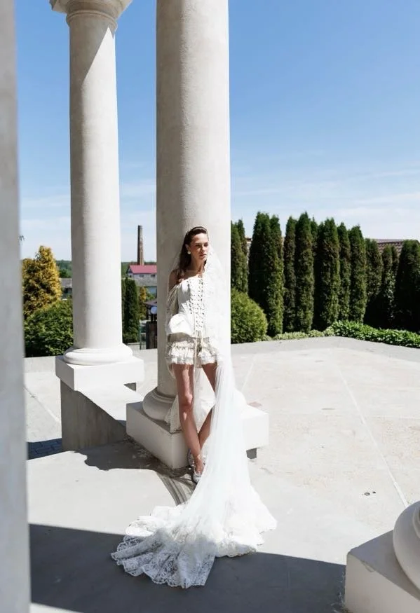 A woman in a white lace wedding dress standing next to classical white columns outdoors with a landscaped background.