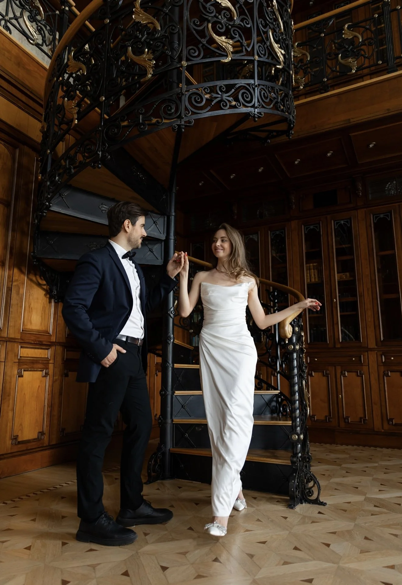 A man in a black tuxedo and woman in a white wedding dress stand inside a wood-paneled room with a spiral staircase. They are smiling at each other, holding hands, with the man touching the staircase. The room features an ornate metal railing and woo