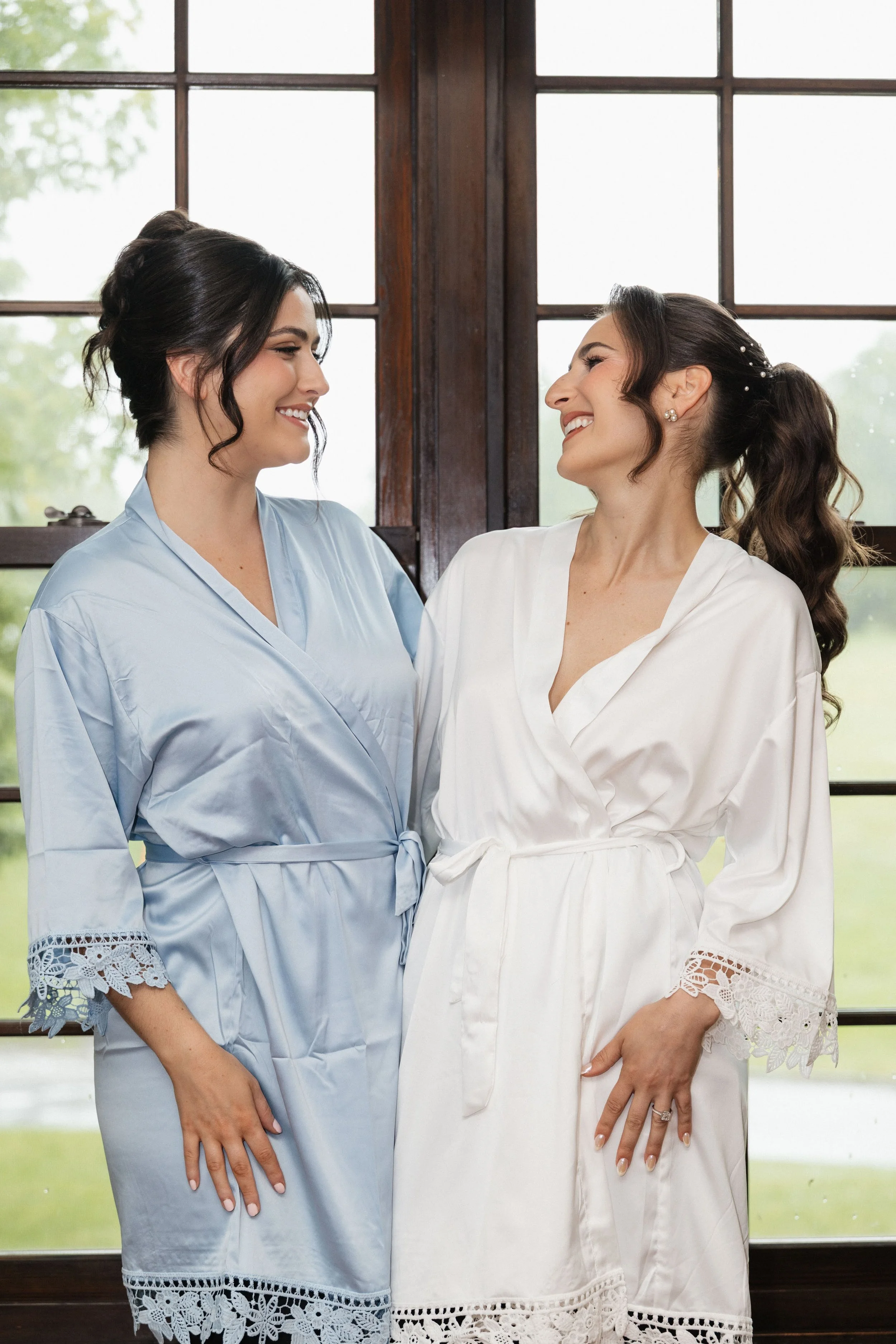 Two women dressed in white and light blue robes smiling at each other indoors near large windows with a green outdoor view.