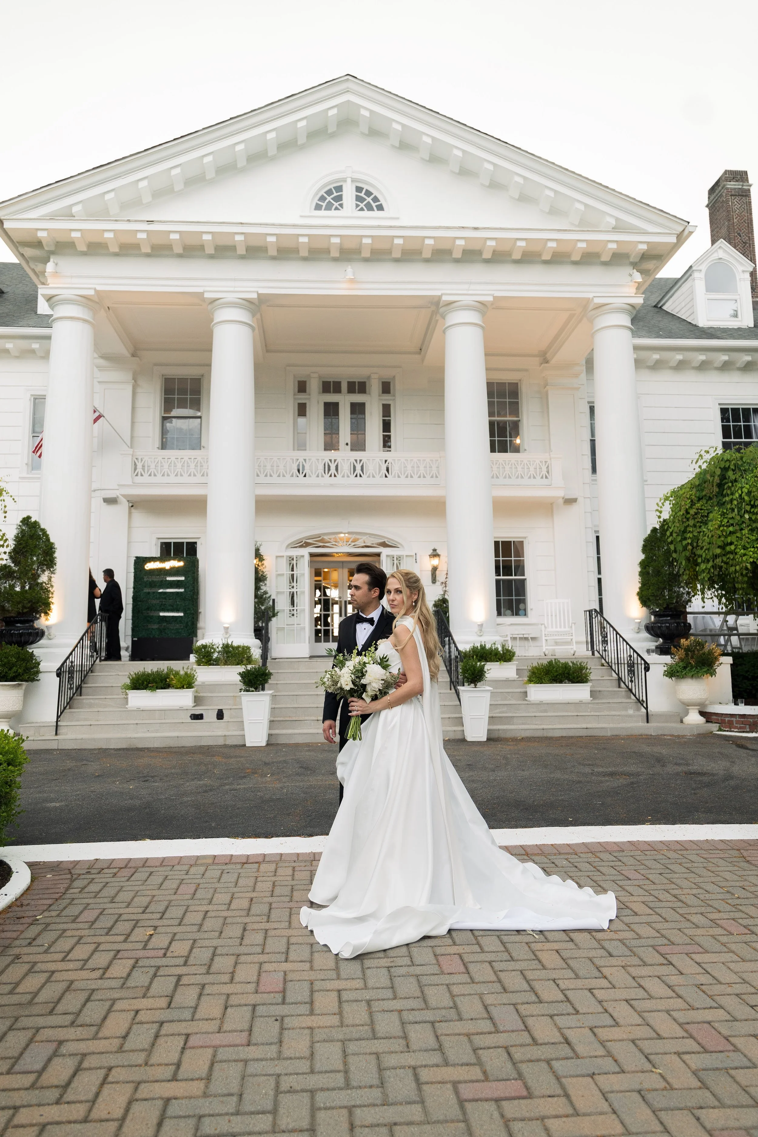 A newlywed couple dressed in wedding attire standing in front of a large white mansion with columns and a balcony, celebrating their wedding outside.