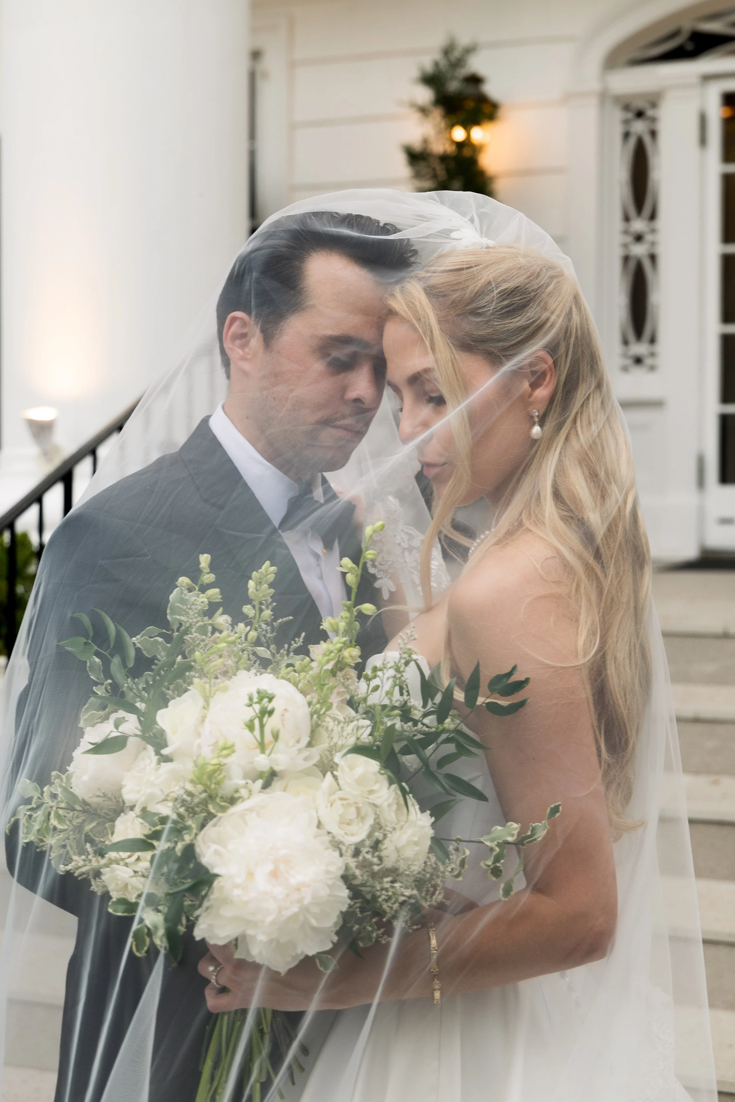 A bride and groom embracing on their wedding day, with the bride wearing a white dress and the groom in a grey suit. They are under a veil, holding a bouquet of white flowers, and standing outdoors in front of a white building.