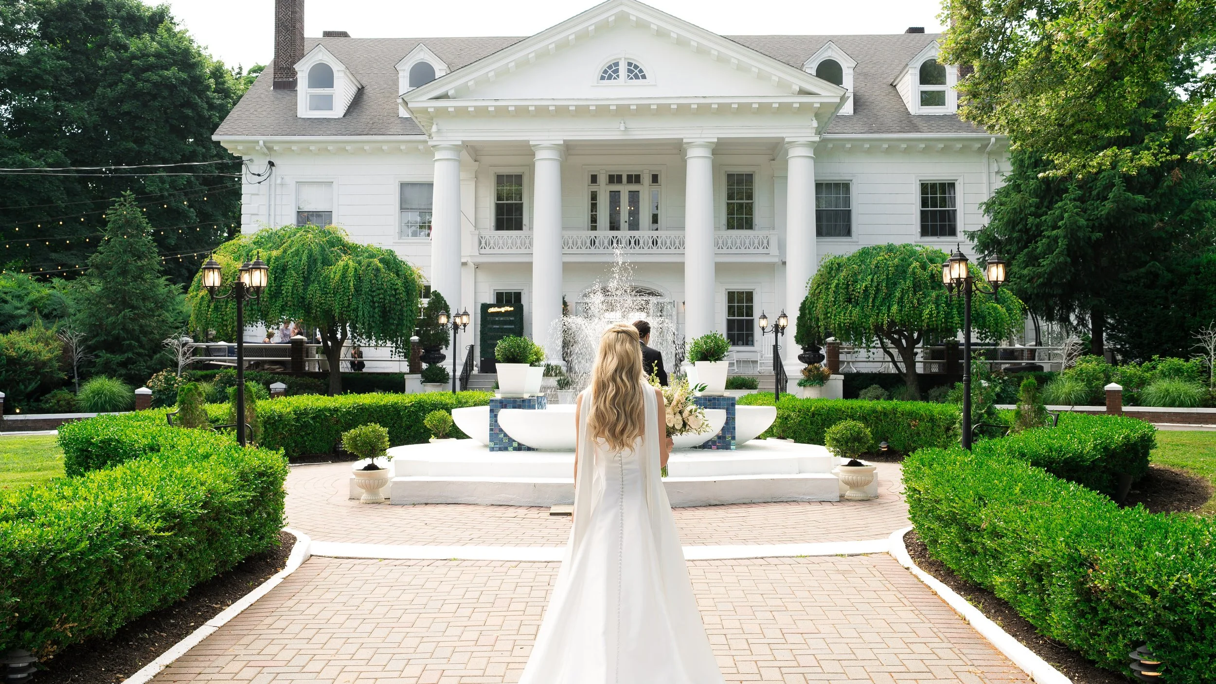 A woman in a white dress with long wavy blonde hair standing in front of a fountain outside a large white mansion with tall columns and a balcony. The garden is well-maintained with trimmed bushes, trees, and black street lamps.