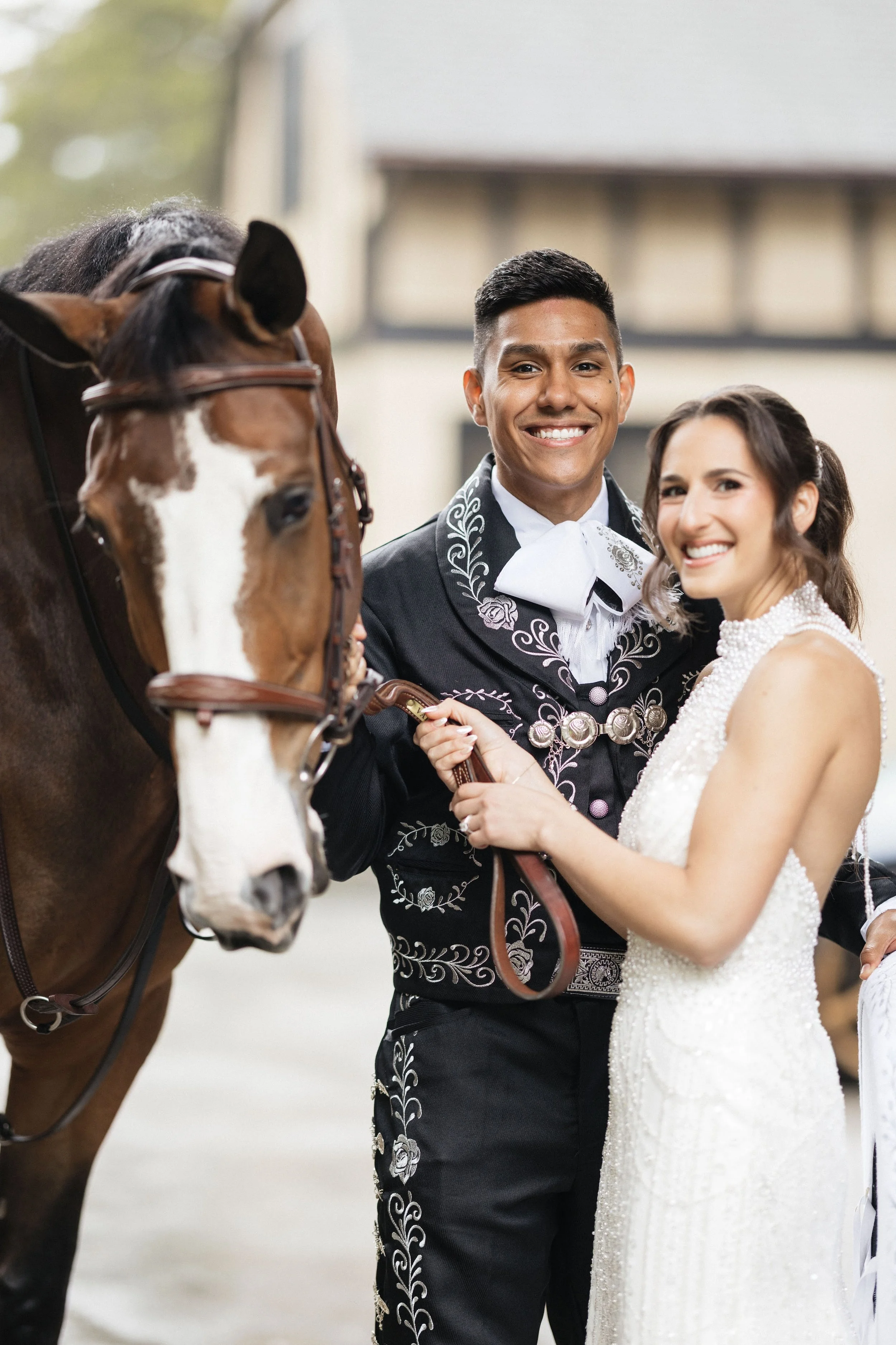 A smiling couple in wedding attire with a brown horse, holding the horse's reins, outdoors on a bright day.