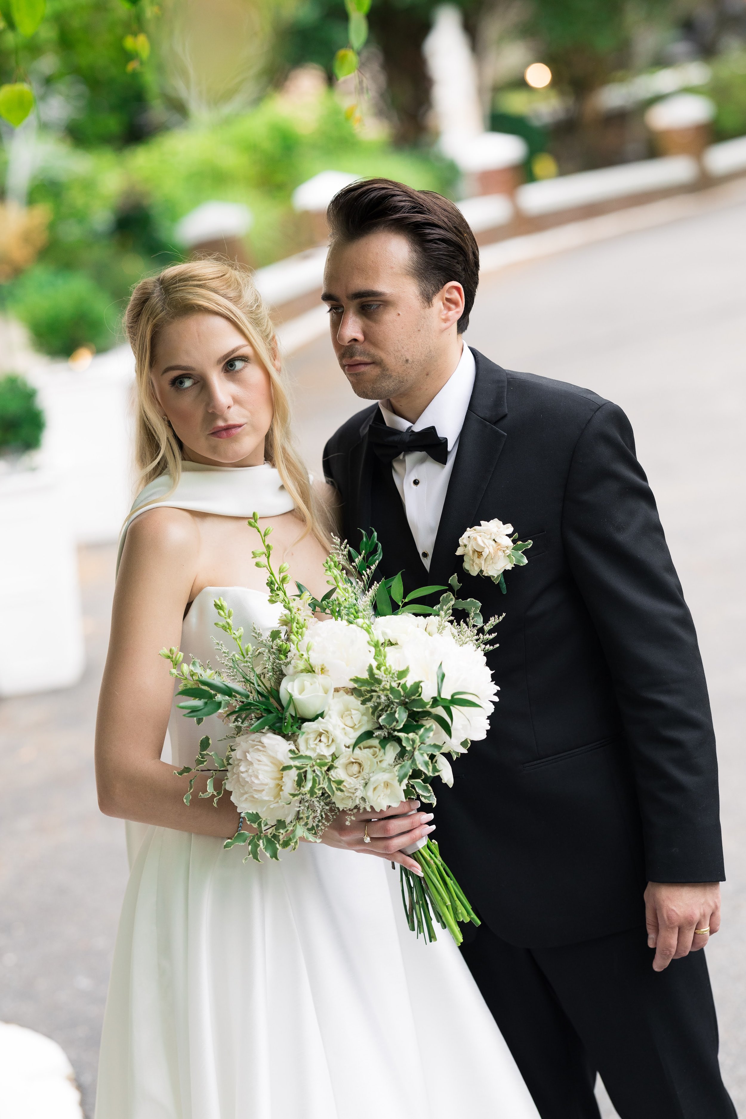 A bride and groom standing close together outdoors, the bride holding a bouquet of white flowers and greenery, both dressed in formal wedding attire.