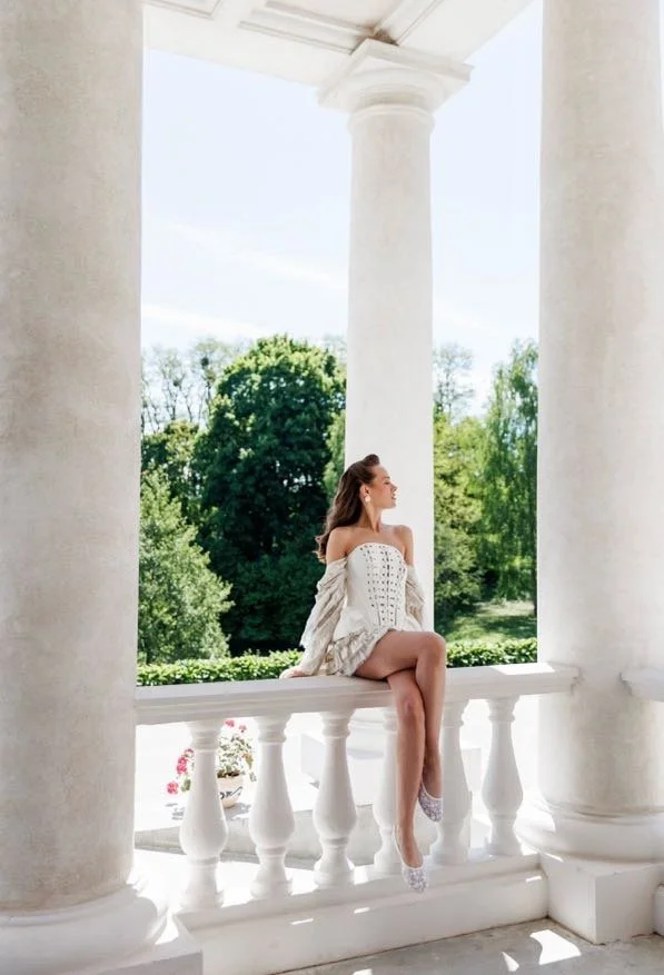 A woman sitting on a white porch railing with large white columns, greenery, and trees in the background.