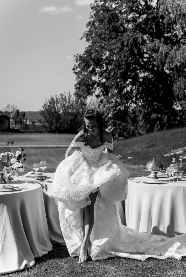 A woman in a wedding dress playfully lifts her skirt as she steps over a table set for a formal outdoor event, with round tables, floral arrangements, and a large tree in the background.