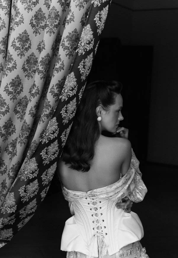 A woman with dark, vintage-style hair and pearl earrings, sitting on a bed, with her back partly to the camera. She is wearing an elegant off-the-shoulder top or dress with lace-up corset detail at her back. She is gazing thoughtfully to the side, pa