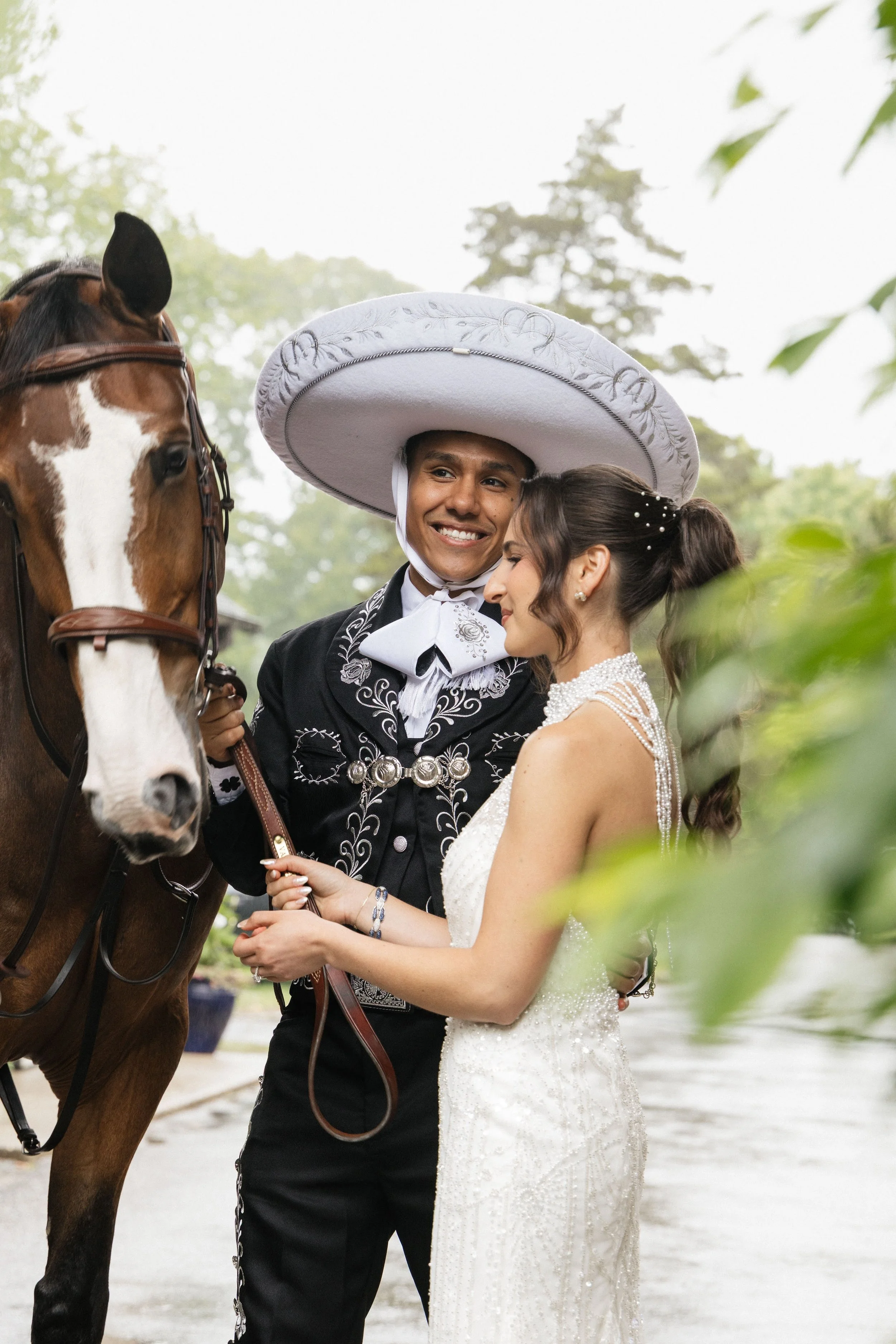 A man wearing a large white sombrero and traditional Mexican clothing, and a woman in a white wedding dress, are standing next to a brown and white horse. They are outdoors, surrounded by trees and greenery, and appear to be sharing a happy moment.