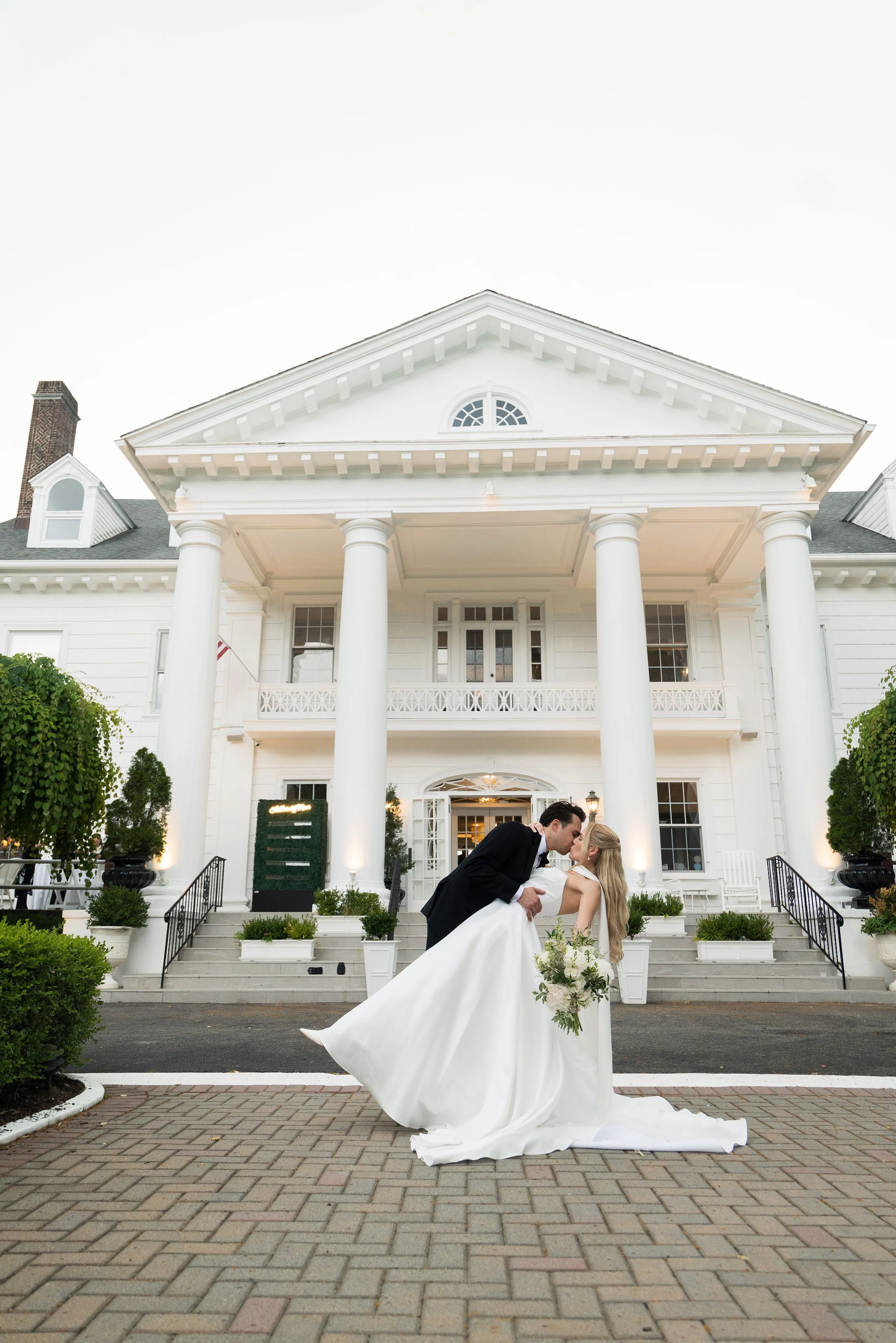 A wedding couple sharing a kiss in front of a white house with columns and a large porch.