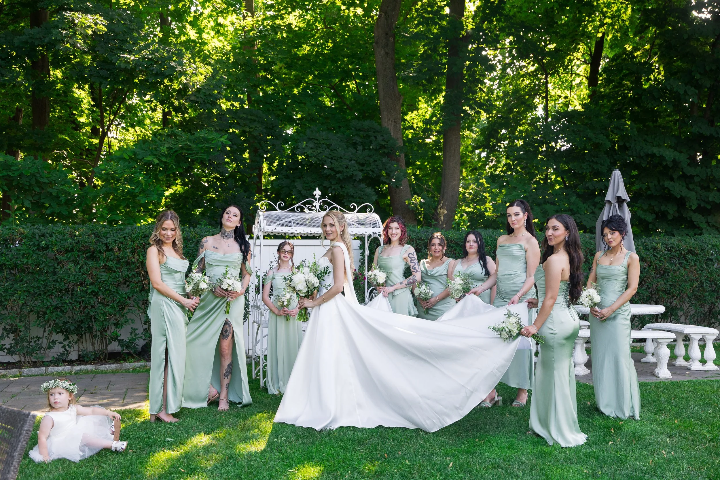 A group of women dressed in matching mint green dresses and a young girl in a white dress standing outdoors in front of a lush green forest, with some holding bouquets, during a wedding celebration.