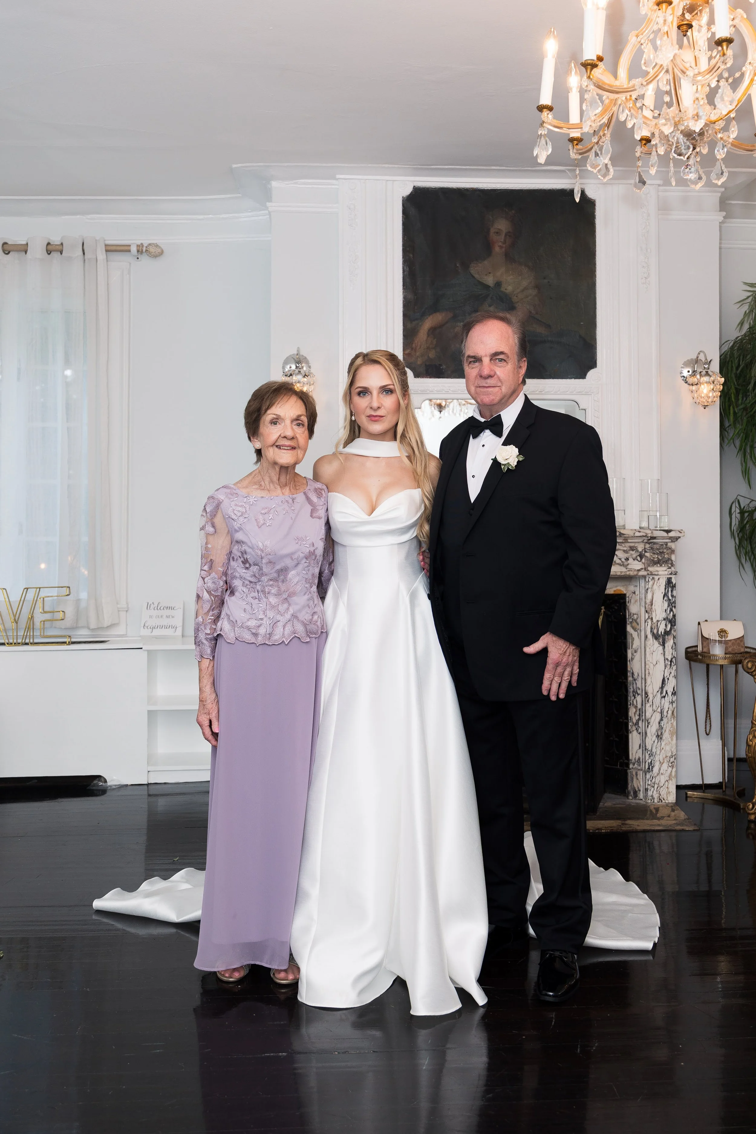 Three people dressed in formal attire standing in a decorated room with a chandelier, fireplace, and painting. The person in the middle is a woman in a white dress, flanked by an elderly woman in a lavender dress and an older man in a black tuxedo.