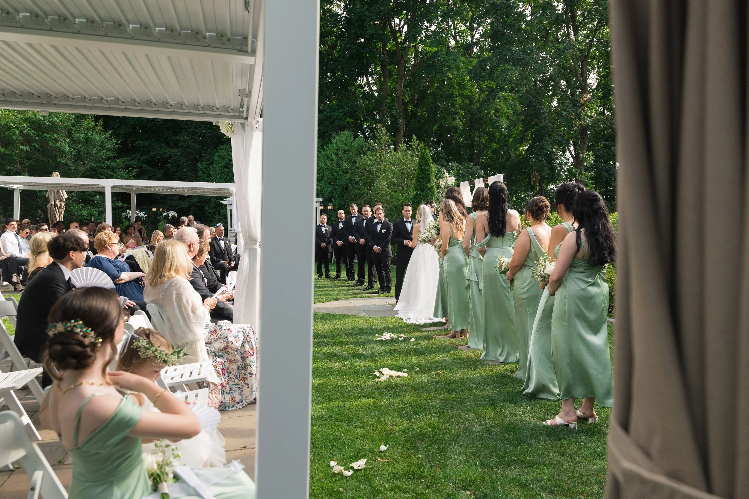 Outdoor wedding ceremony with guests seated on white chairs, bridesmaids in light green dresses, and groomsmen in black tuxedos, set in a garden with tall trees and lush greenery.