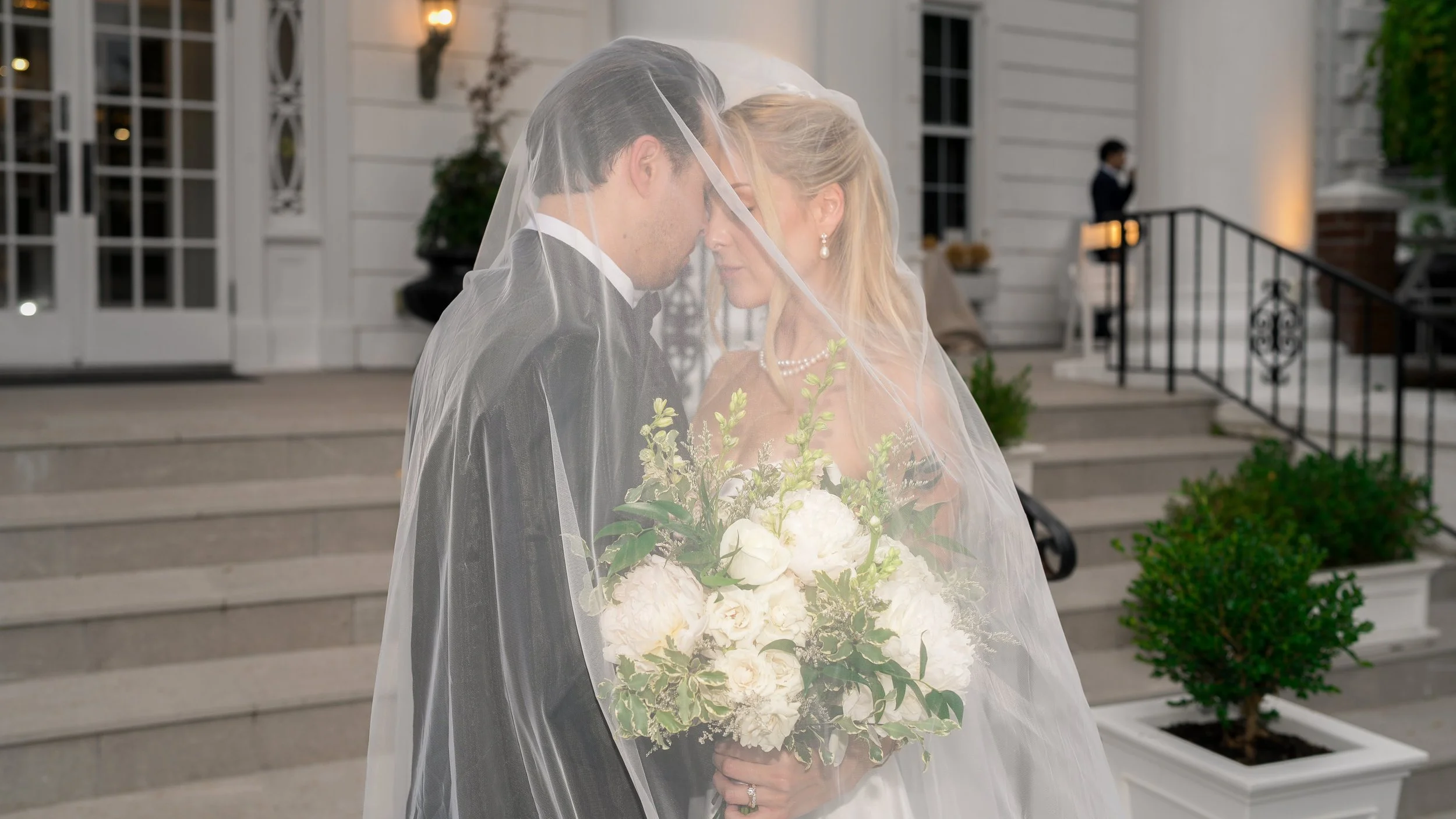 A bride and groom under a veil, close together, standing on front steps of a building, with a woman in the background near the door holding a drink, and potted plants on each side of the stairs.