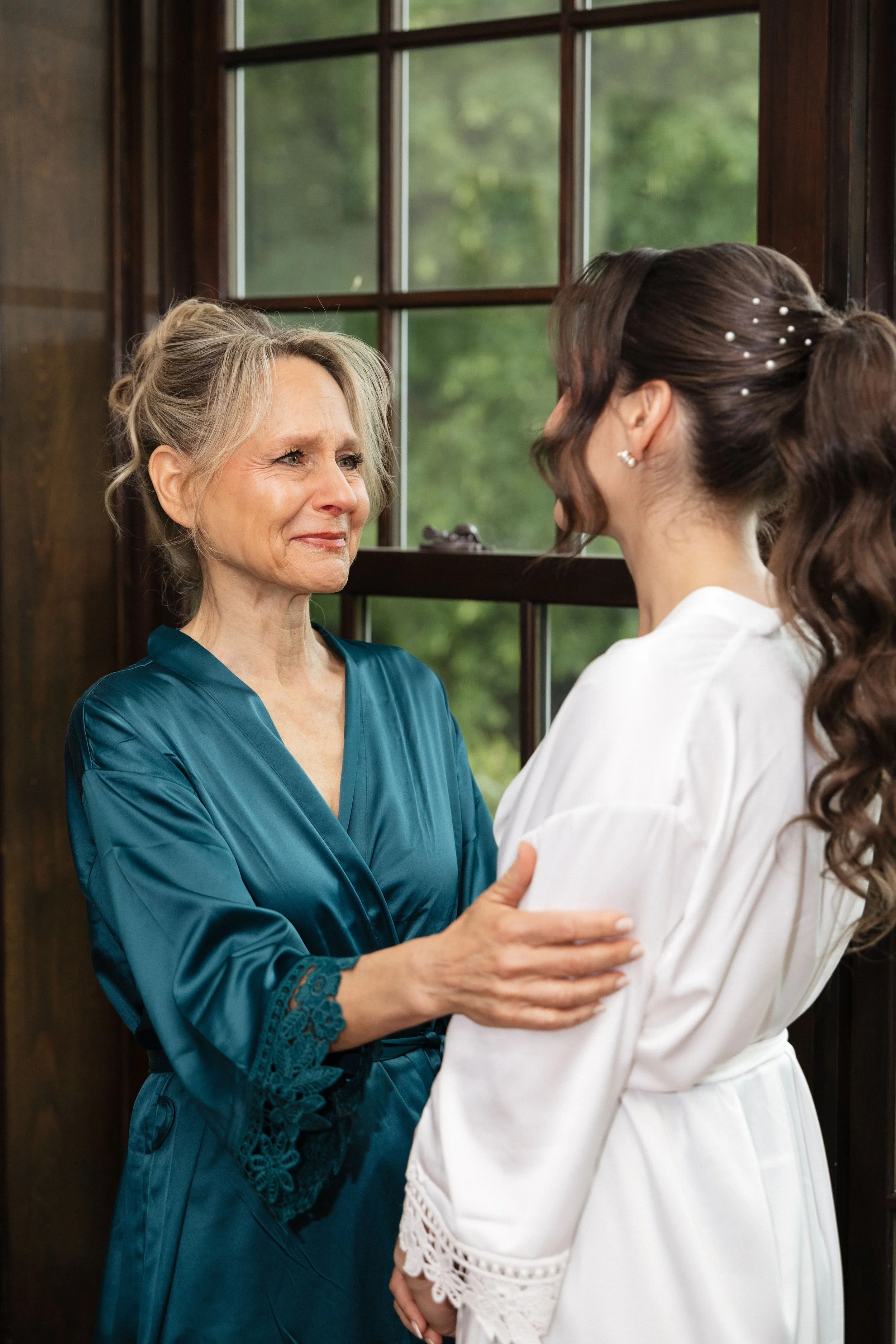 An elderly woman and a younger woman, possibly mother and daughter, sharing an emotional moment indoors by a window. The elderly woman is smiling gently, wearing a teal satin robe with lace details, holding the younger woman's arms. The younger woman