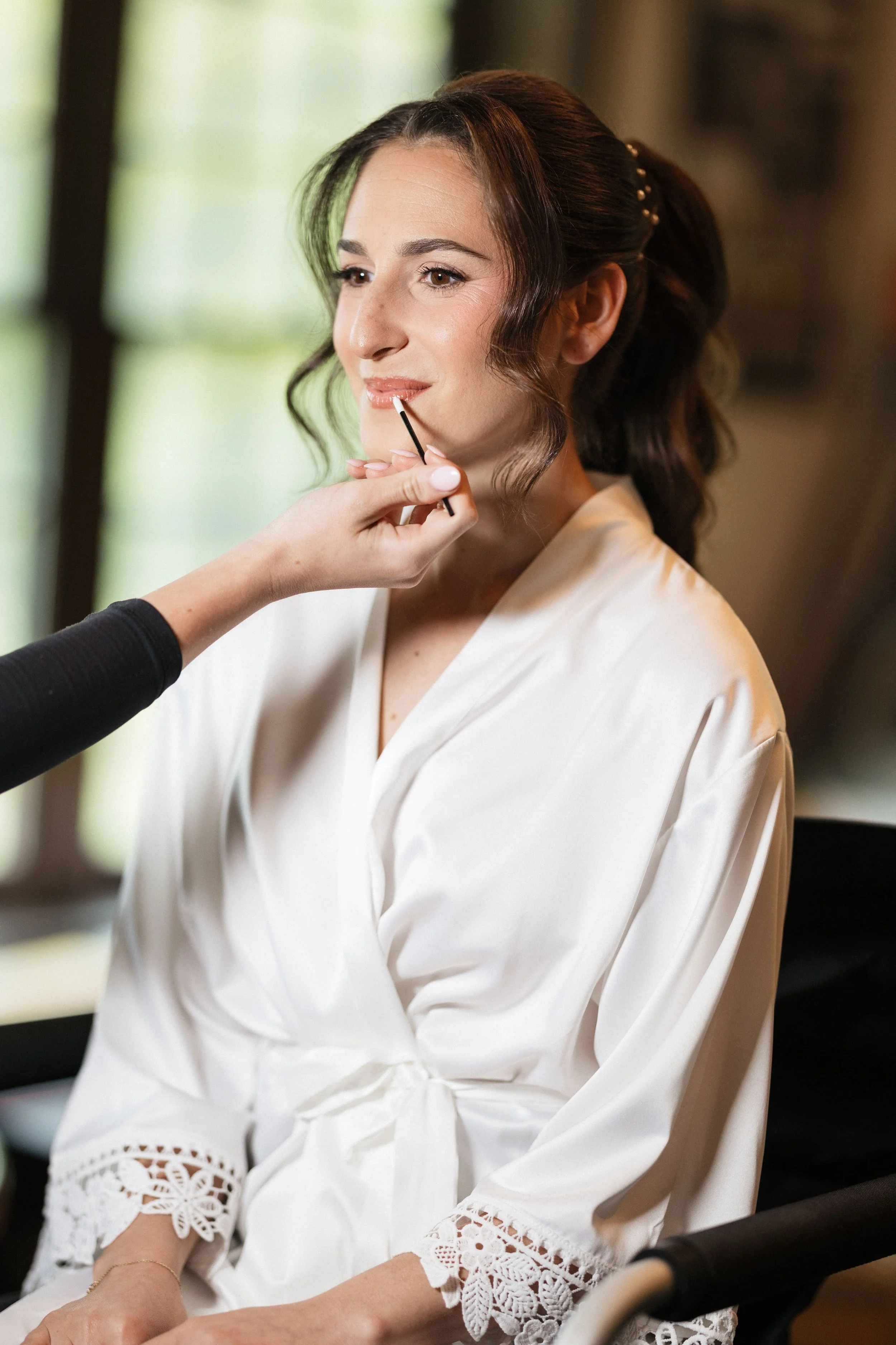 A woman with brown hair styled in loose waves, wearing a white satin robe with lace details on the sleeves, is having her makeup done by a makeup artist who is applying lip gloss or lipstick.