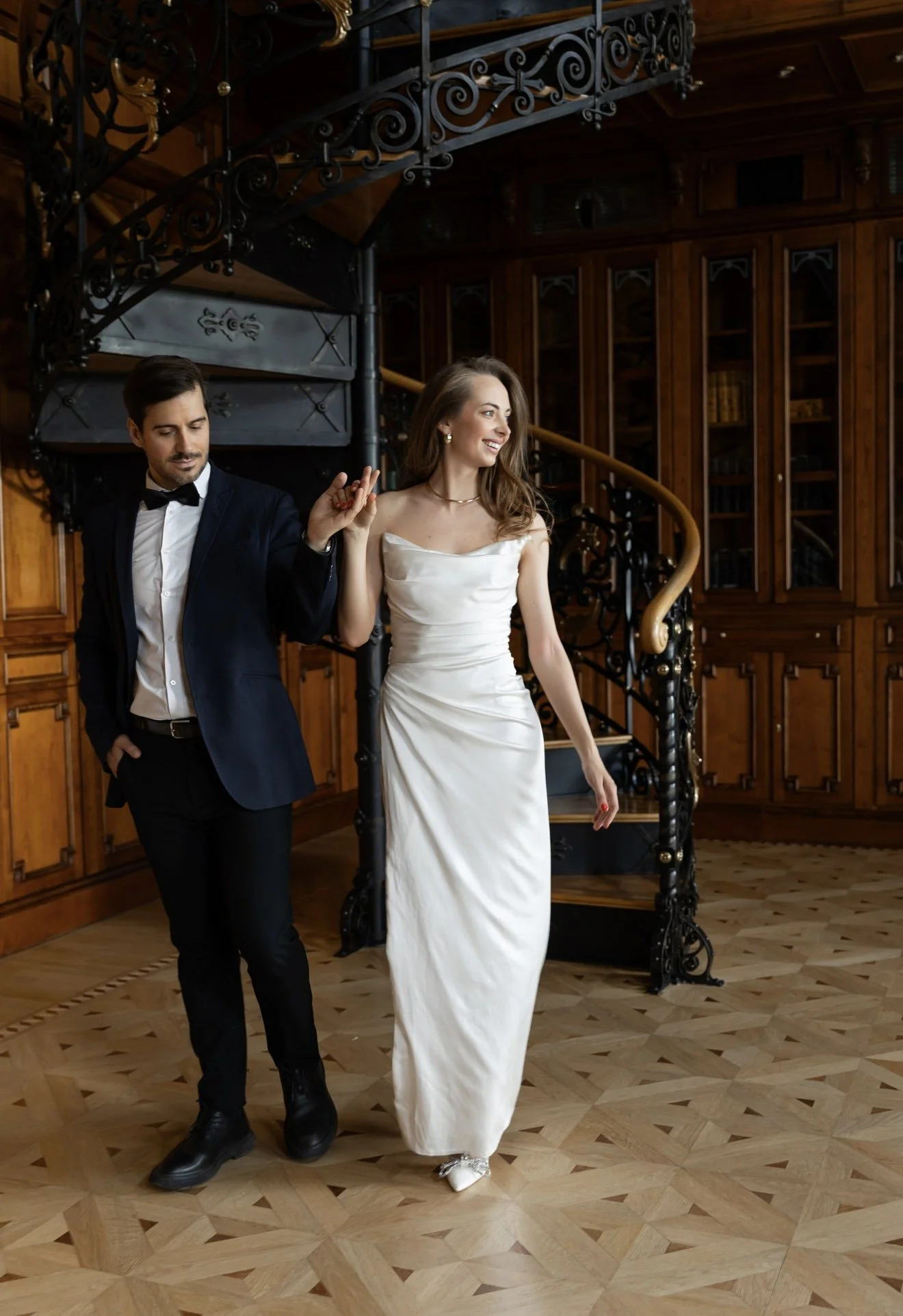 A man in a tuxedo and a woman in a white dress dancing in a polished wooden room with bookshelves and a spiral staircase.