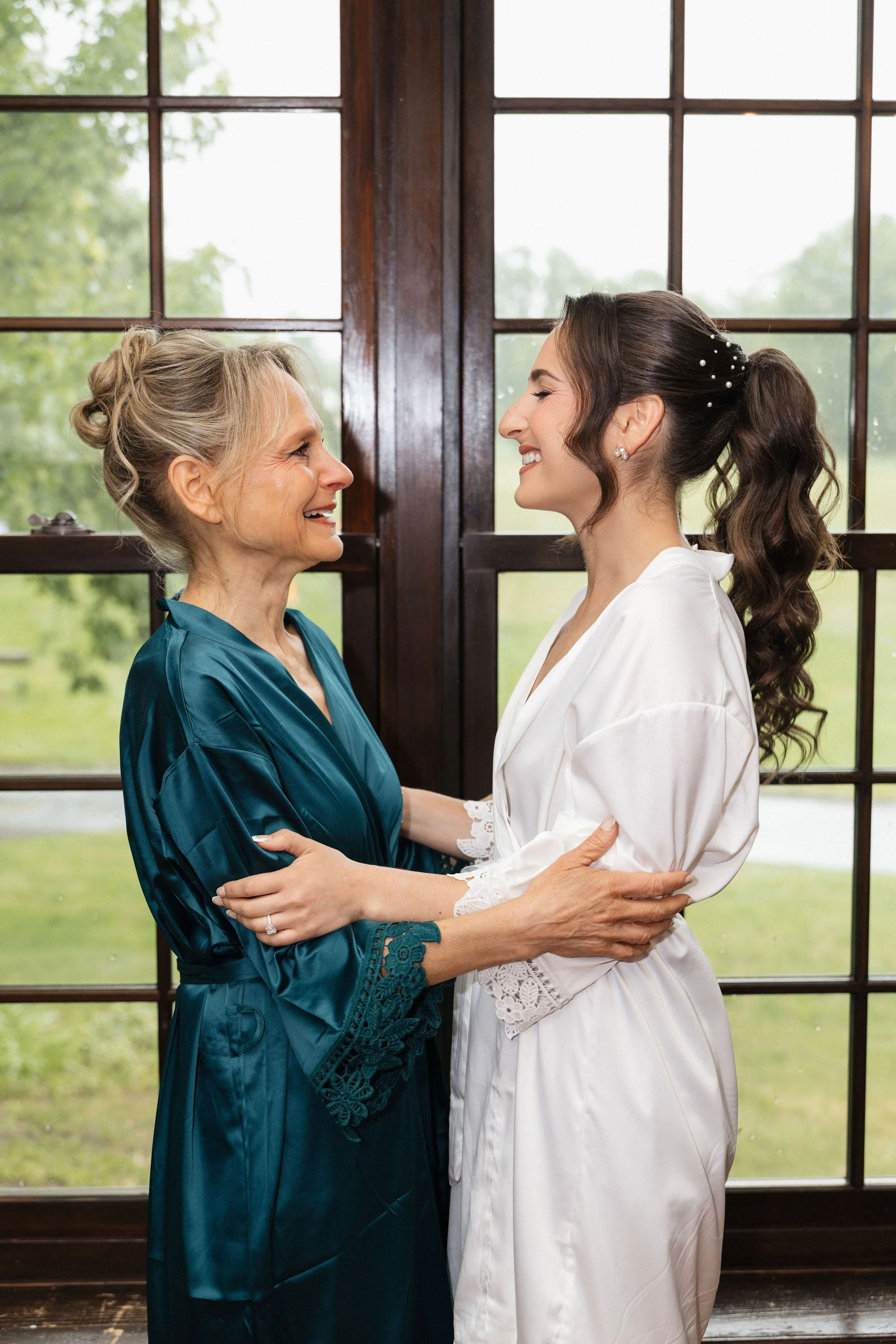 A young woman in a white satin robe with lace trim on the sleeves, smiling and holding hands with an older woman in a teal silk robe, both standing face to face and smiling at each other in front of large windows showing green trees outside.