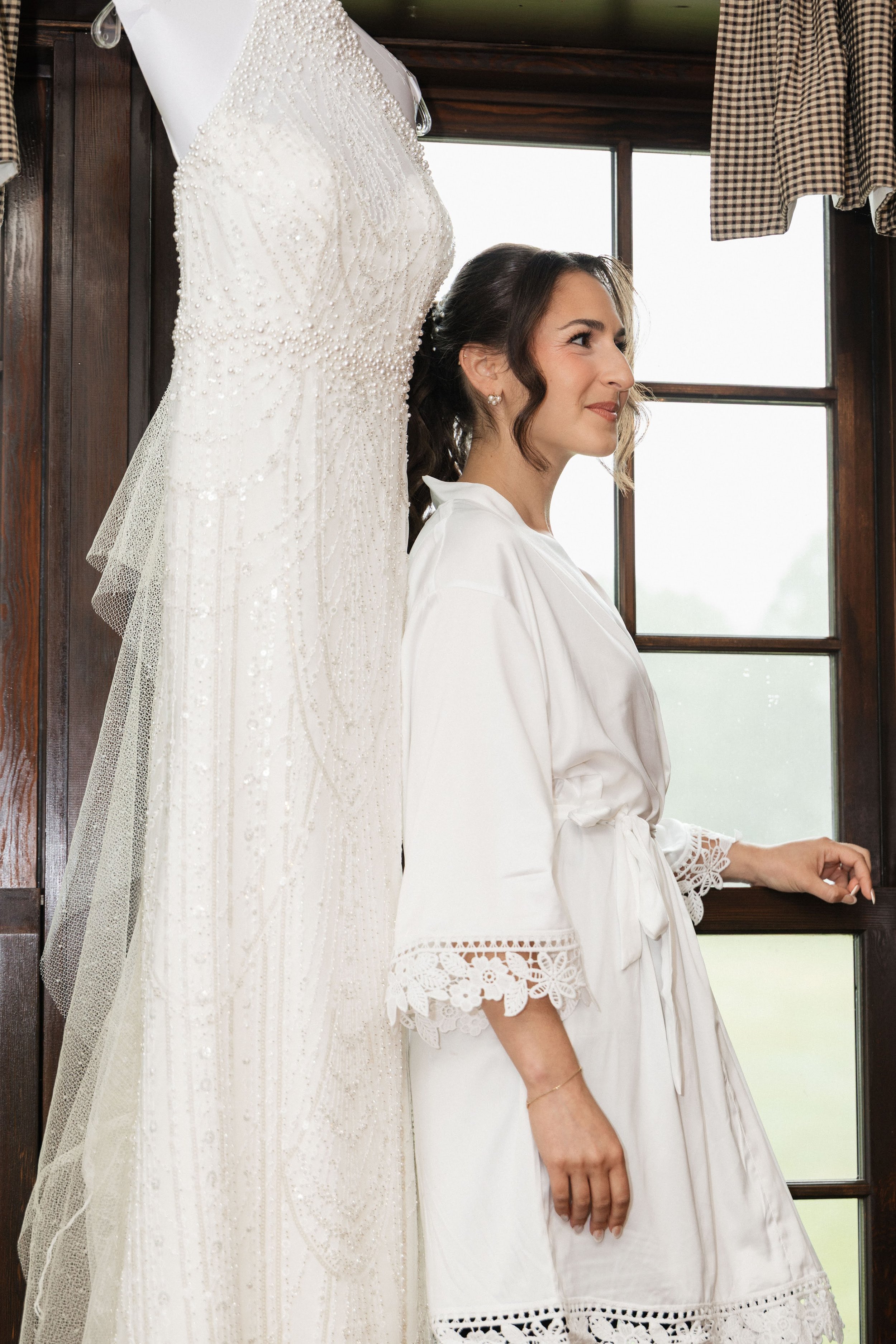 A woman with brown hair styled in loose waves, wearing pearl earrings and a white satin robe with lace details on the sleeves, stands next to a wedding dress hanging on a wooden wall. The wedding dress is white, strapless, and heavily embellished wit