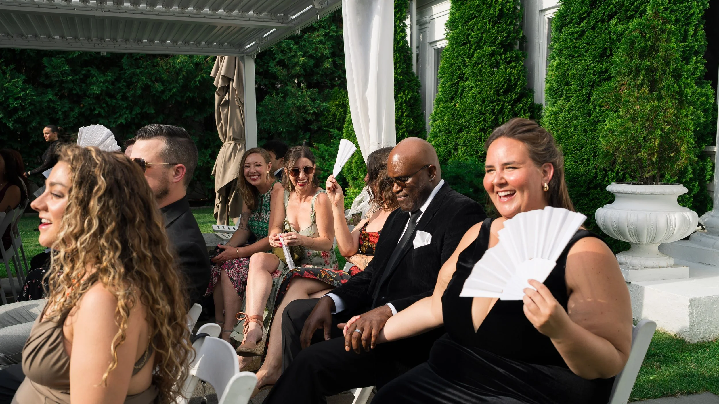 People sitting outdoors at a wedding or celebration, smiling and holding white fans, with green bushes and white decorative urns in the background.