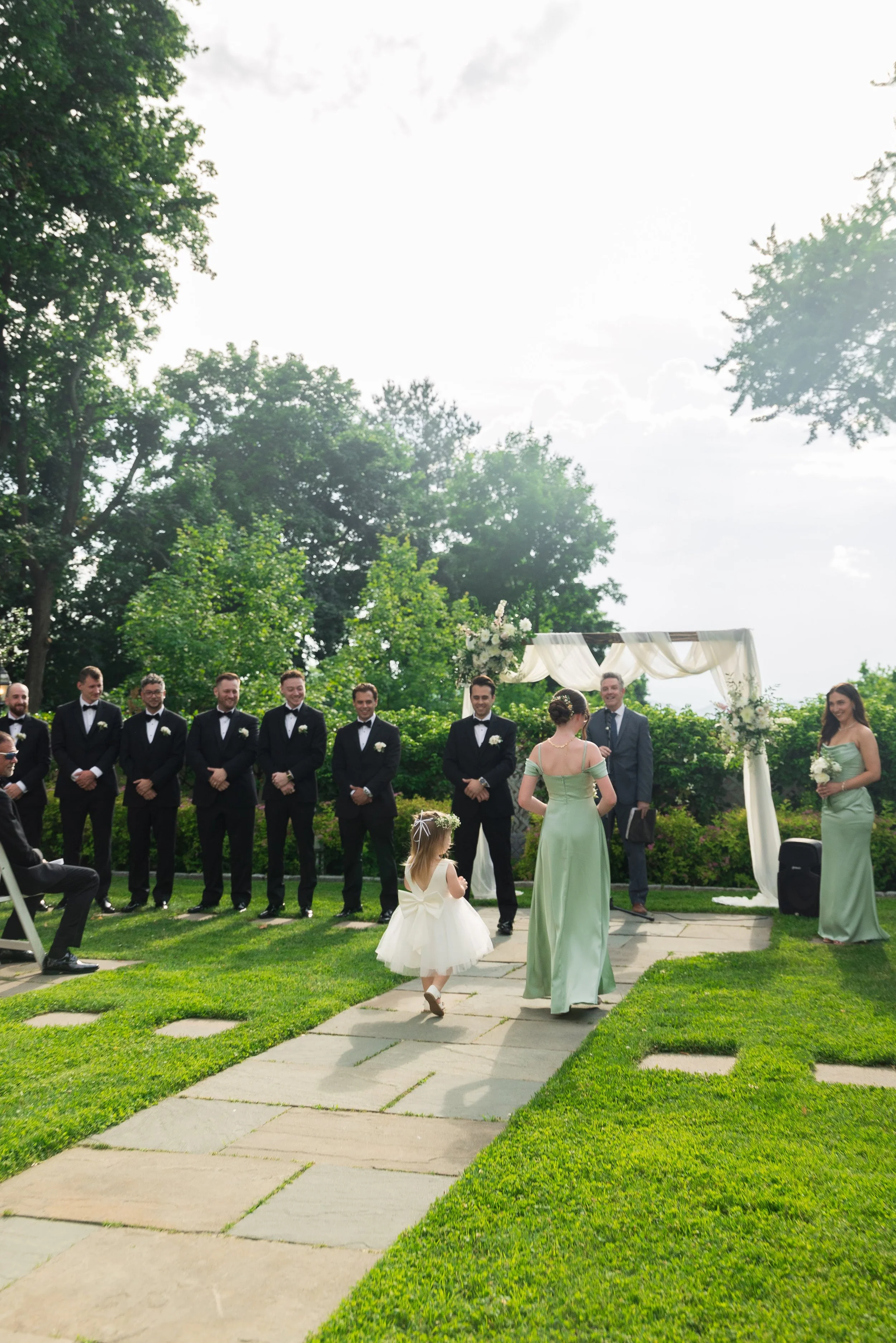A wedding ceremony outdoors with a bride, groom, and bridesmaids, along with groomsmen, standing under a decorated arch. A flower girl walks along a stone path on a lush green lawn, with trees and a cloudy sky in the background.