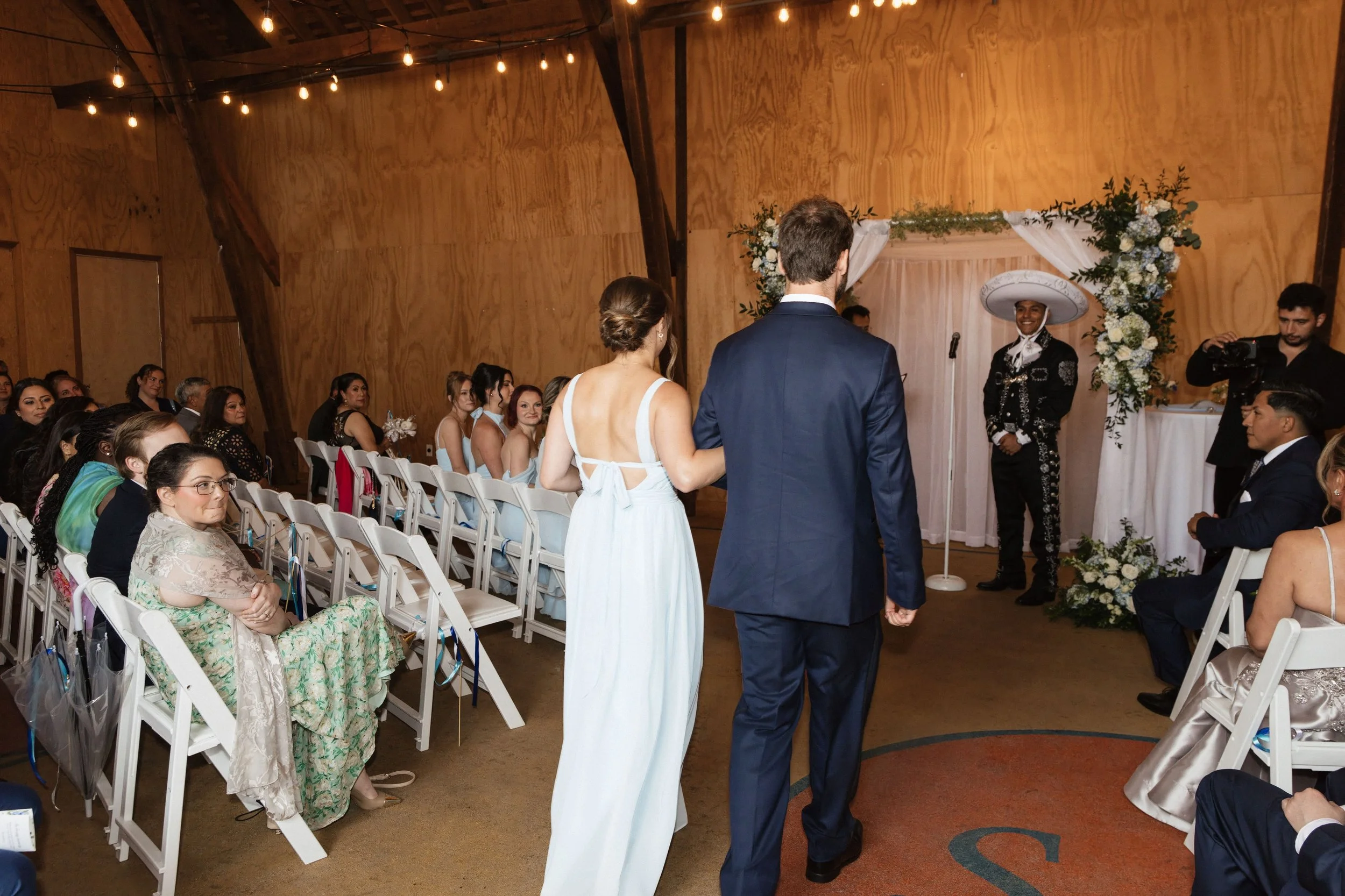 A wedding ceremony taking place in a wooden hall with string lights. The bride and groom stand facing an officiant in front of a floral backdrop. Guests are seated on white chairs, watching the couple.
