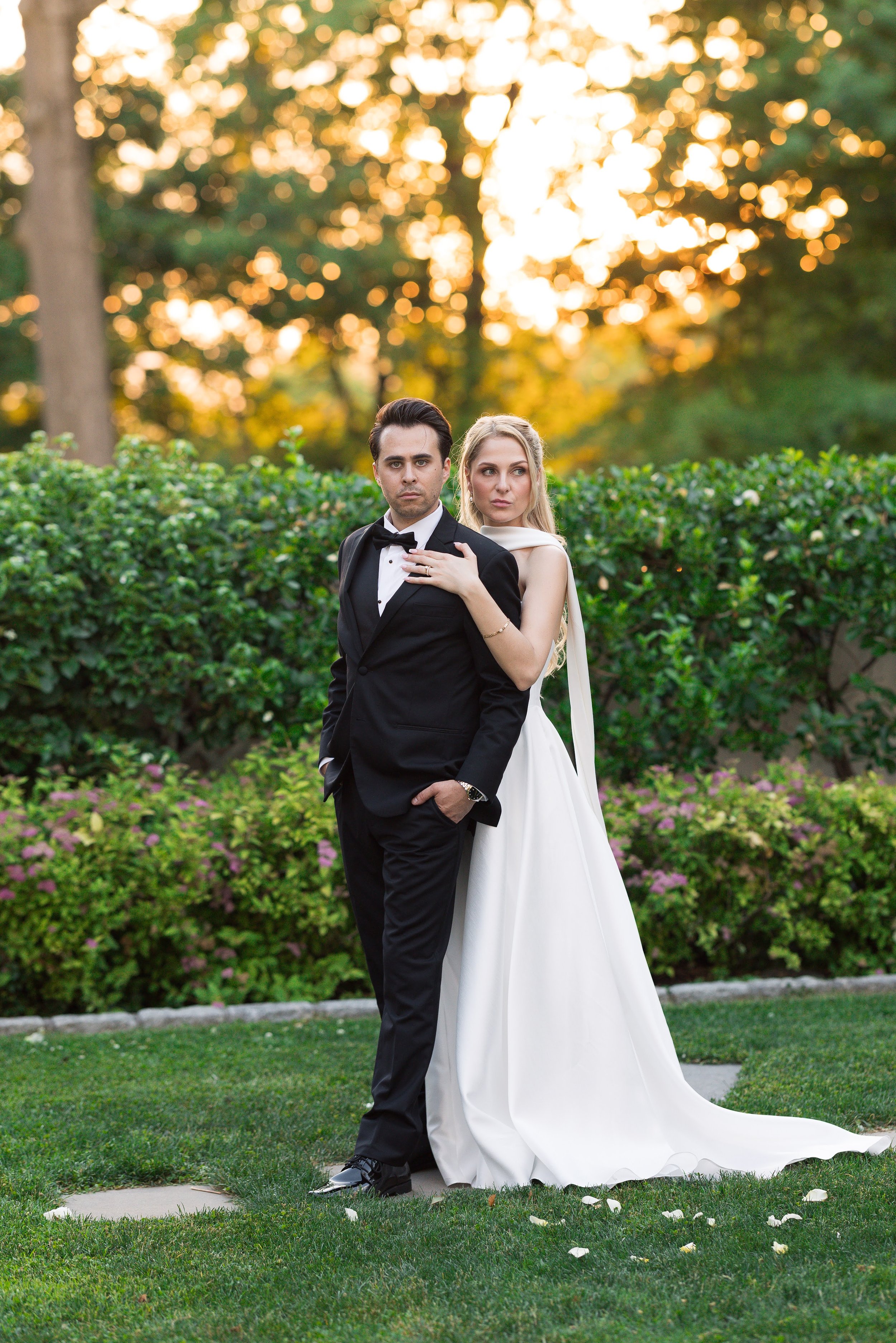 A young man in a black tuxedo and a young woman in a white wedding dress stand outdoors on grass with a background of green bushes and trees. The woman has her hand on the man's shoulder, and they both look off into the distance. The sun is setting, 