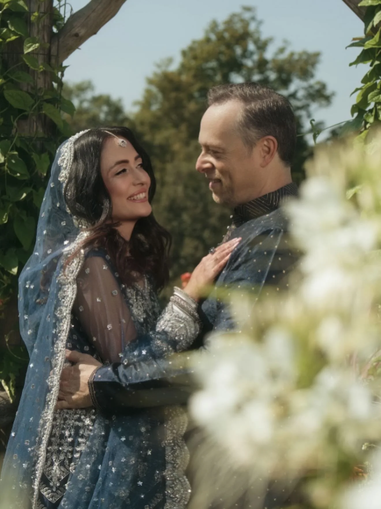 A couple dressed in traditional Indian wedding attire standing close together, smiling, outdoors with trees and a clear sky in the background.